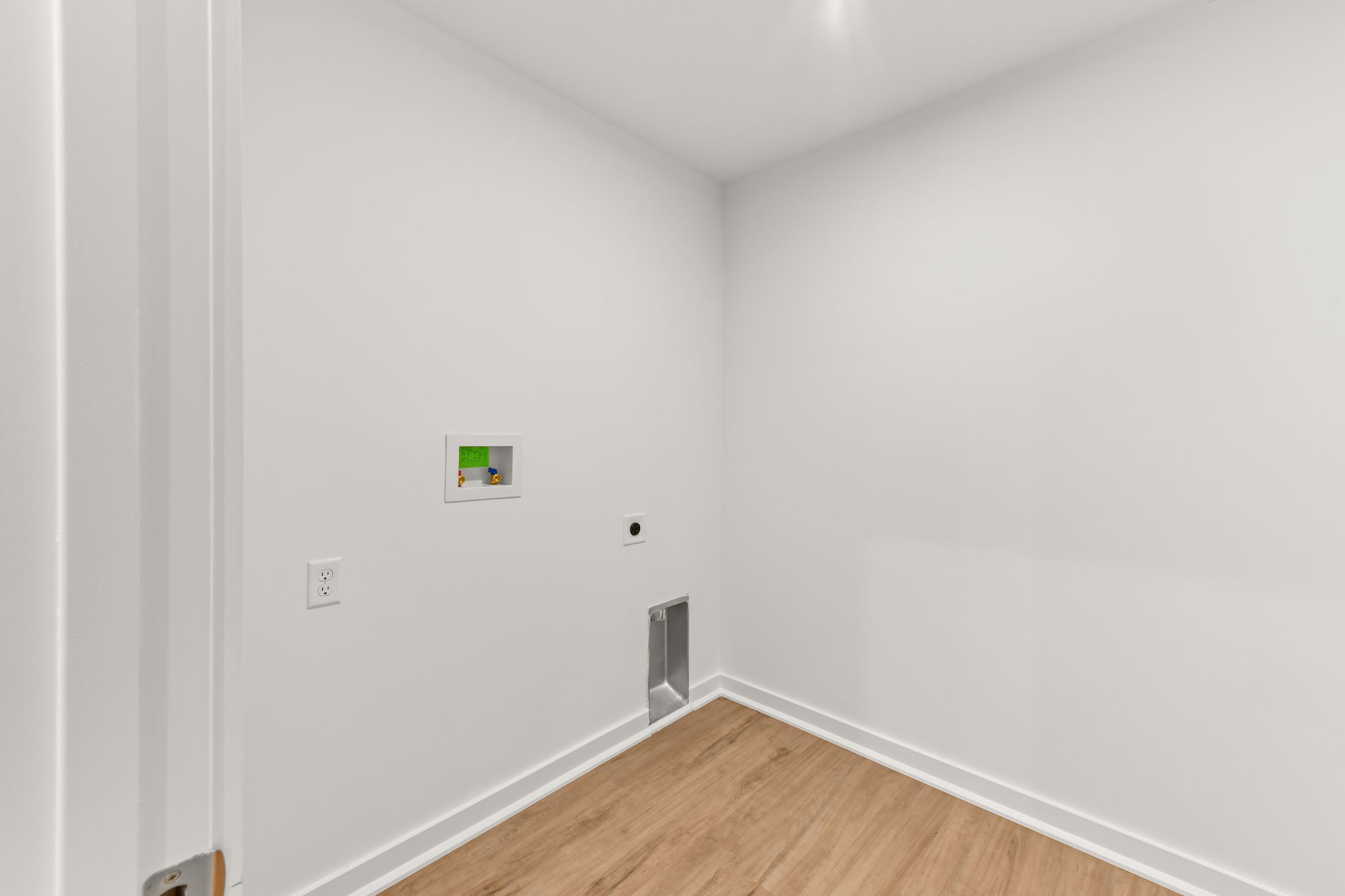 Empty laundry room with white walls, hardwood flooring, plumbing connections, and electrical outlet.