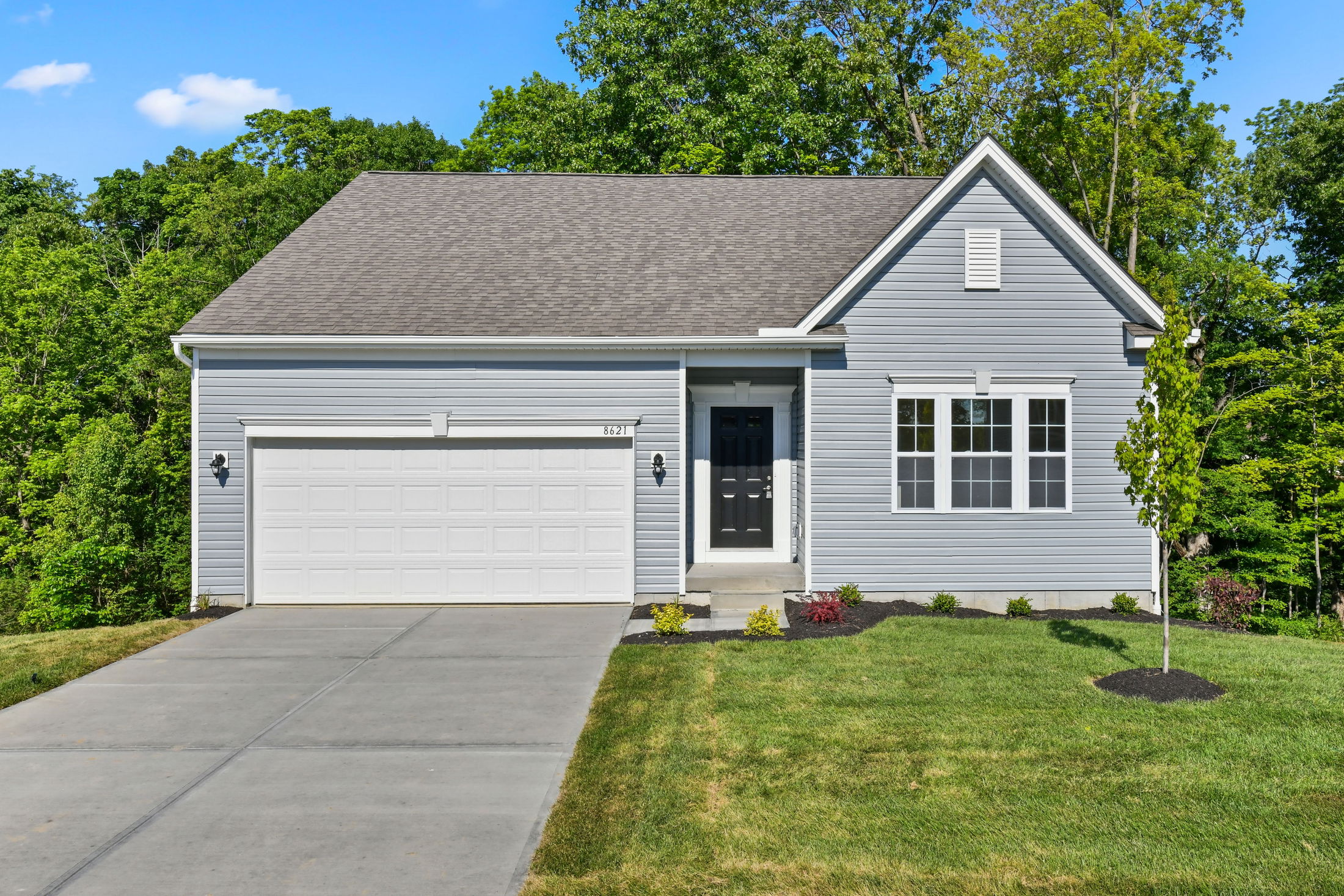 Modern single-story gray house with two-car garage and front yard landscaping against a backdrop of lush green trees.