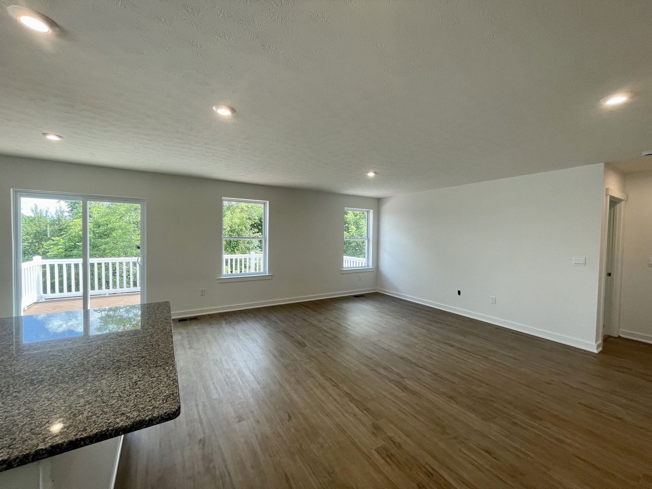Spacious open-concept living room with wooden flooring, large windows, and a kitchen counter, overlooking a greenery-filled balcony.