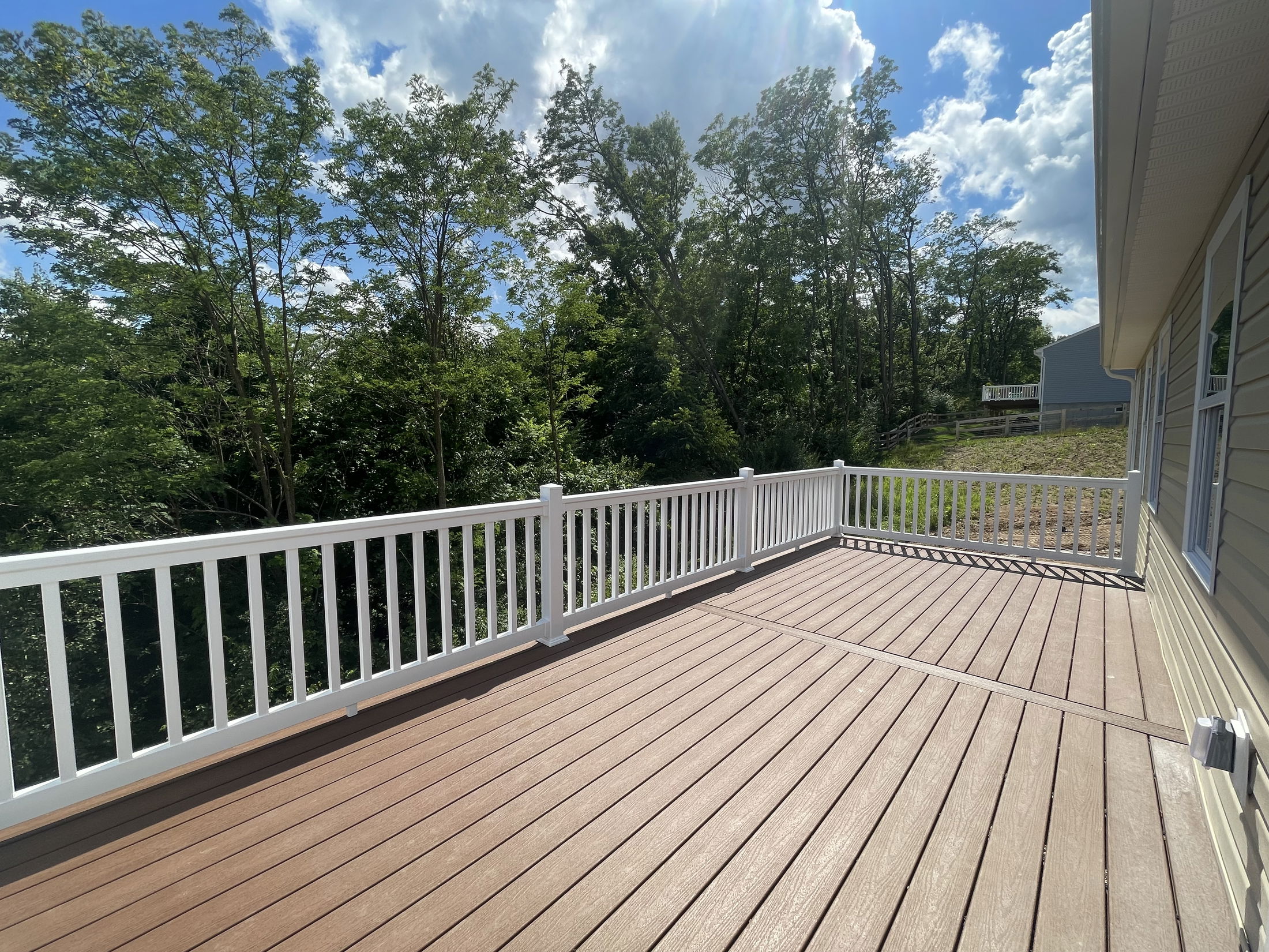 Sunny outdoor deck with white railing overlooking lush green trees and a neighboring house.