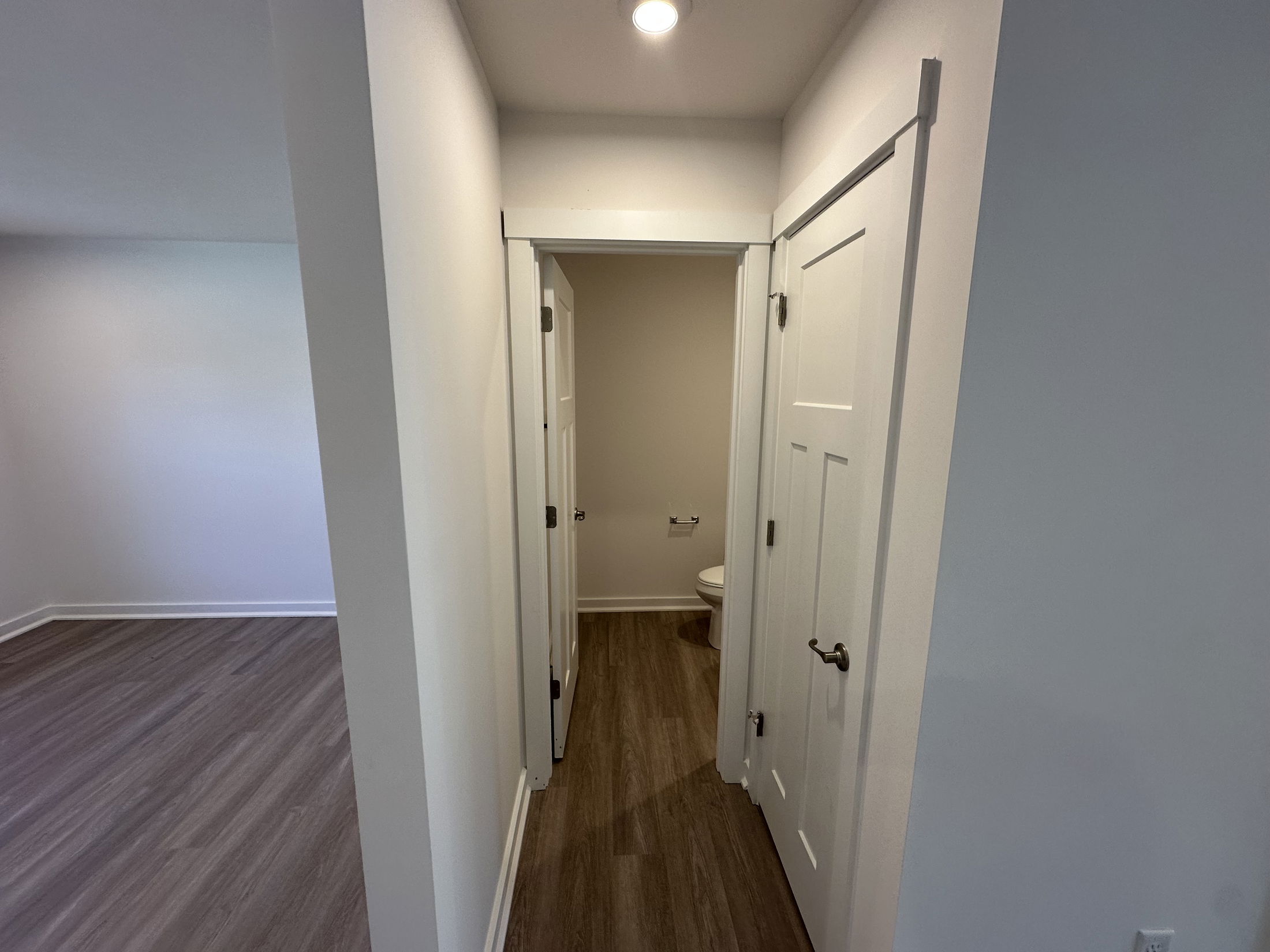 A bright, narrow hallway in a modern home with white doors leads to a bathroom featuring wood-style flooring.