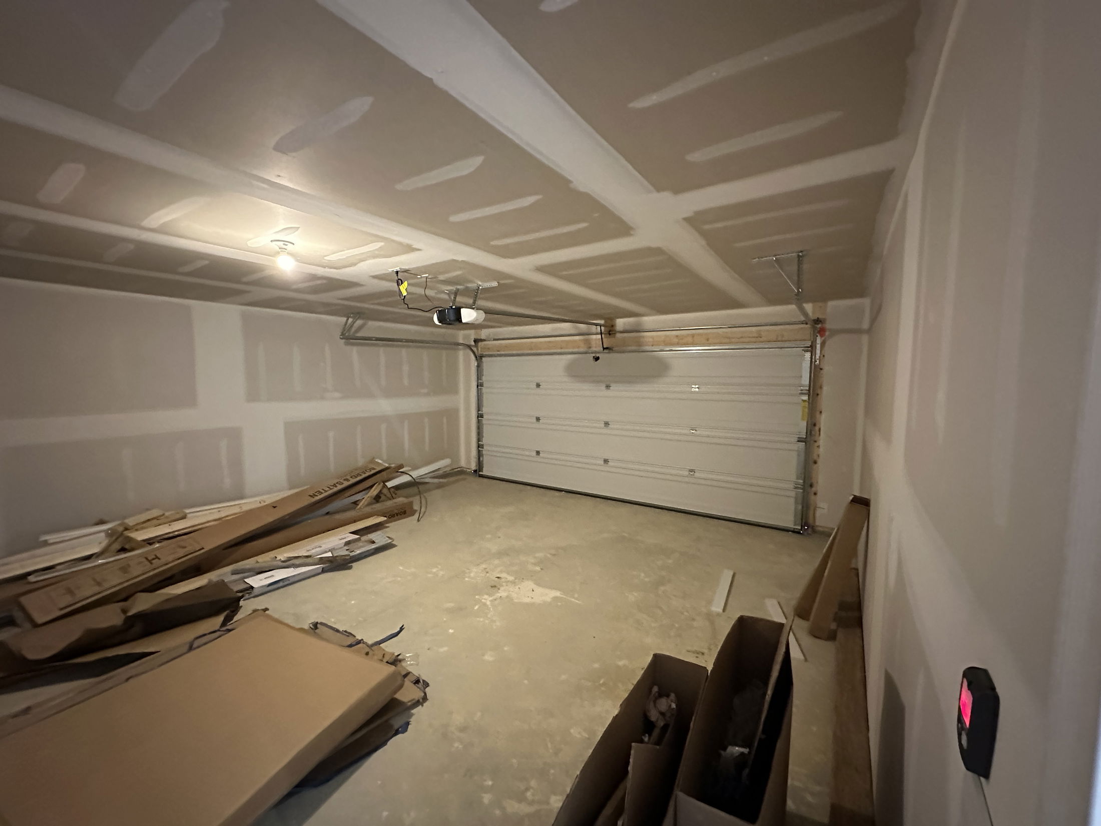 Empty garage with unfinished drywall, scattered construction materials, and an overhead door opener.