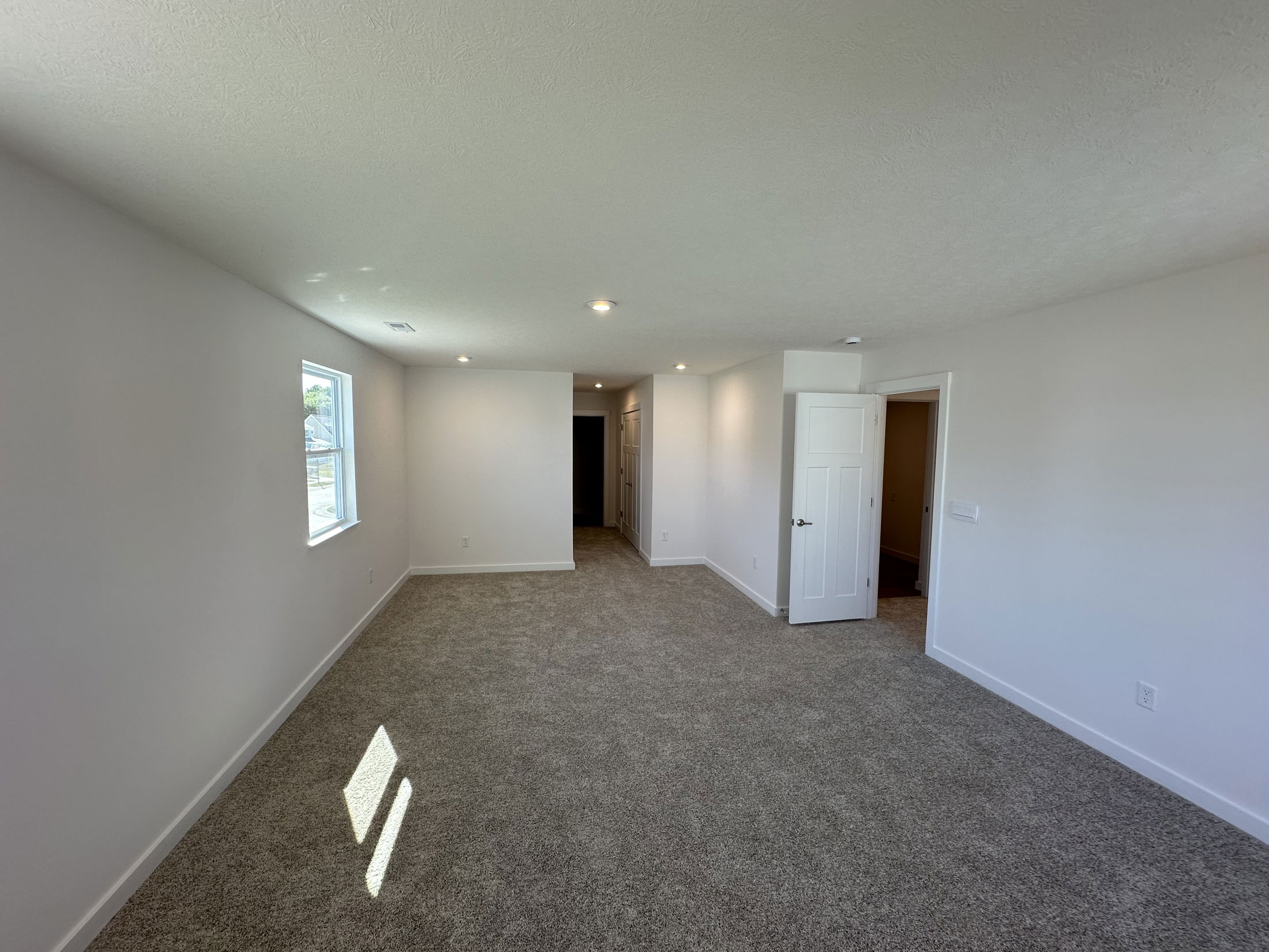 Spacious, newly renovated room with beige carpeting, white walls, recessed lighting, open door, and a large window.