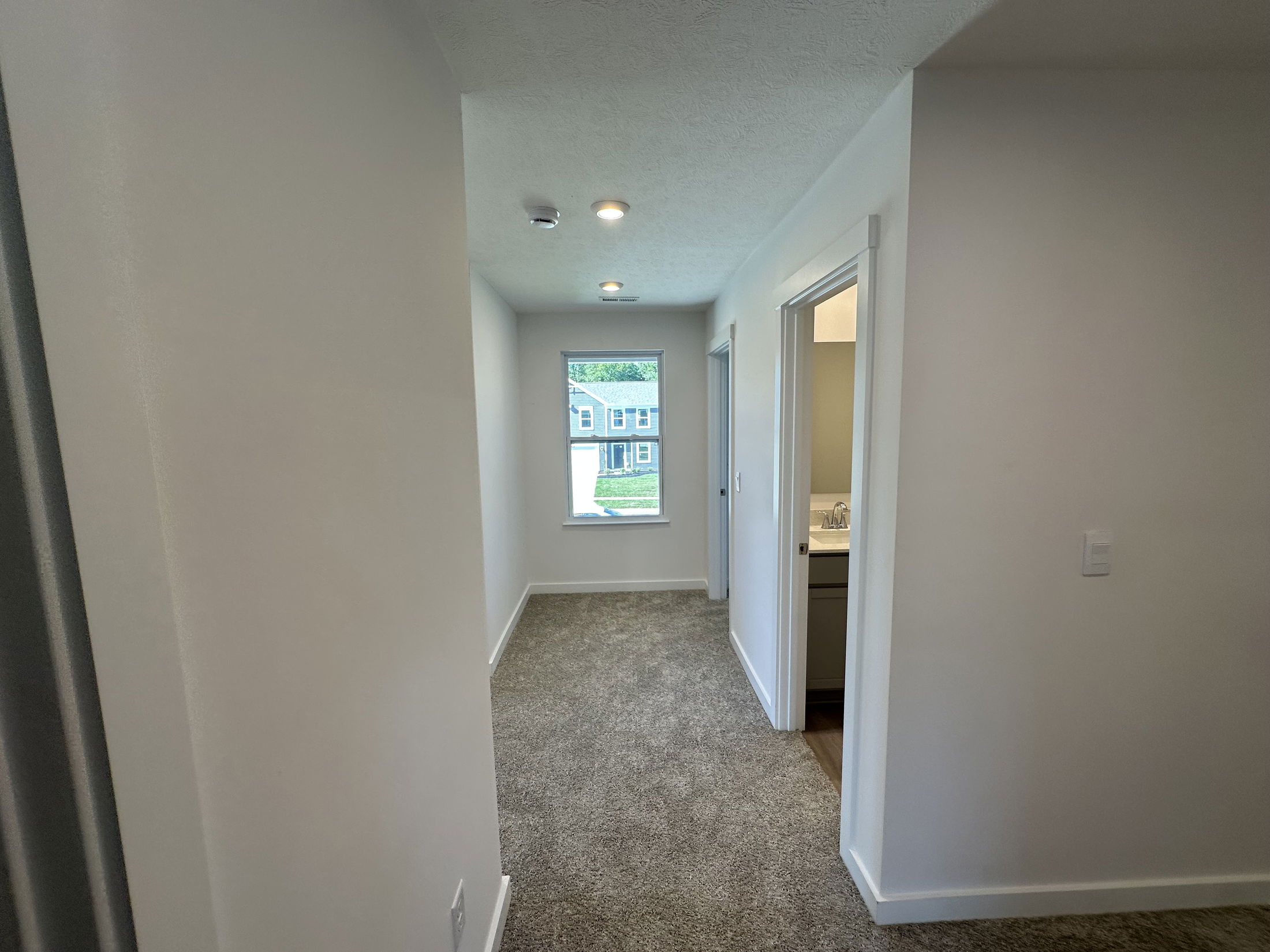 A carpeted hallway in a modern home with white walls and lit ceiling, featuring a window and bathroom entrance.