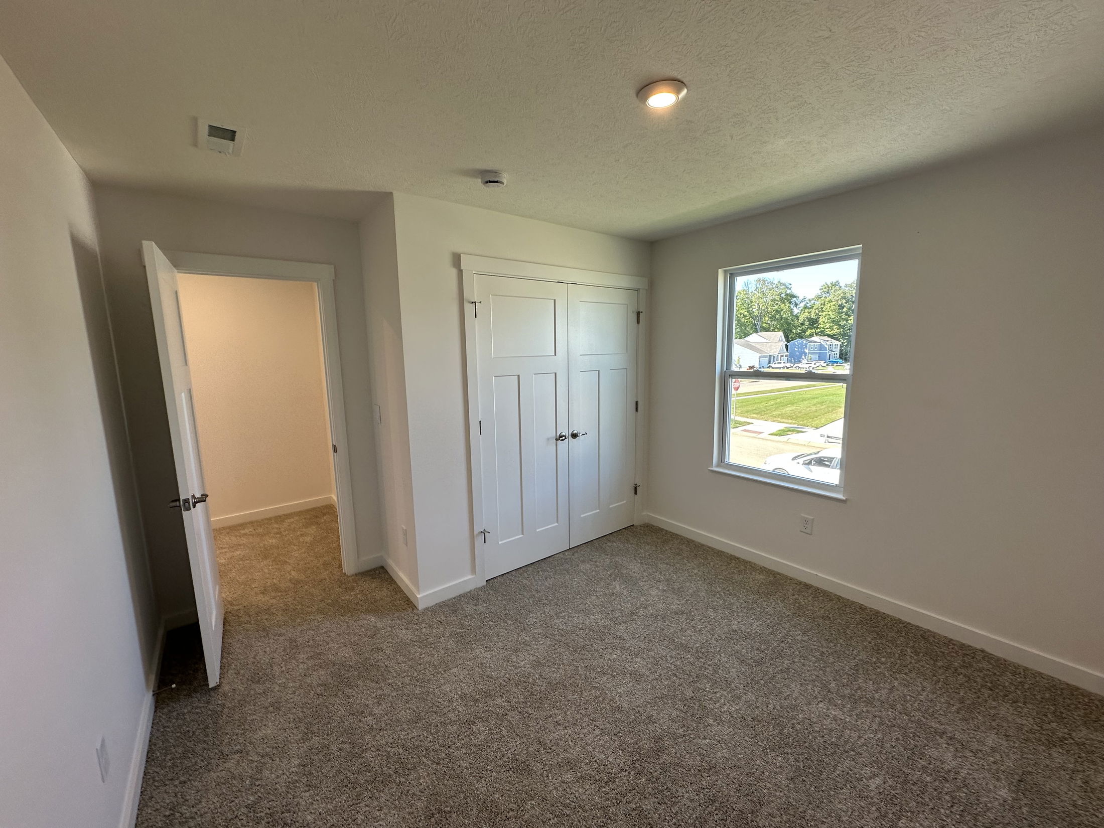 Bright, empty bedroom with plush carpet, white closet doors, and a window overlooking a suburban street.