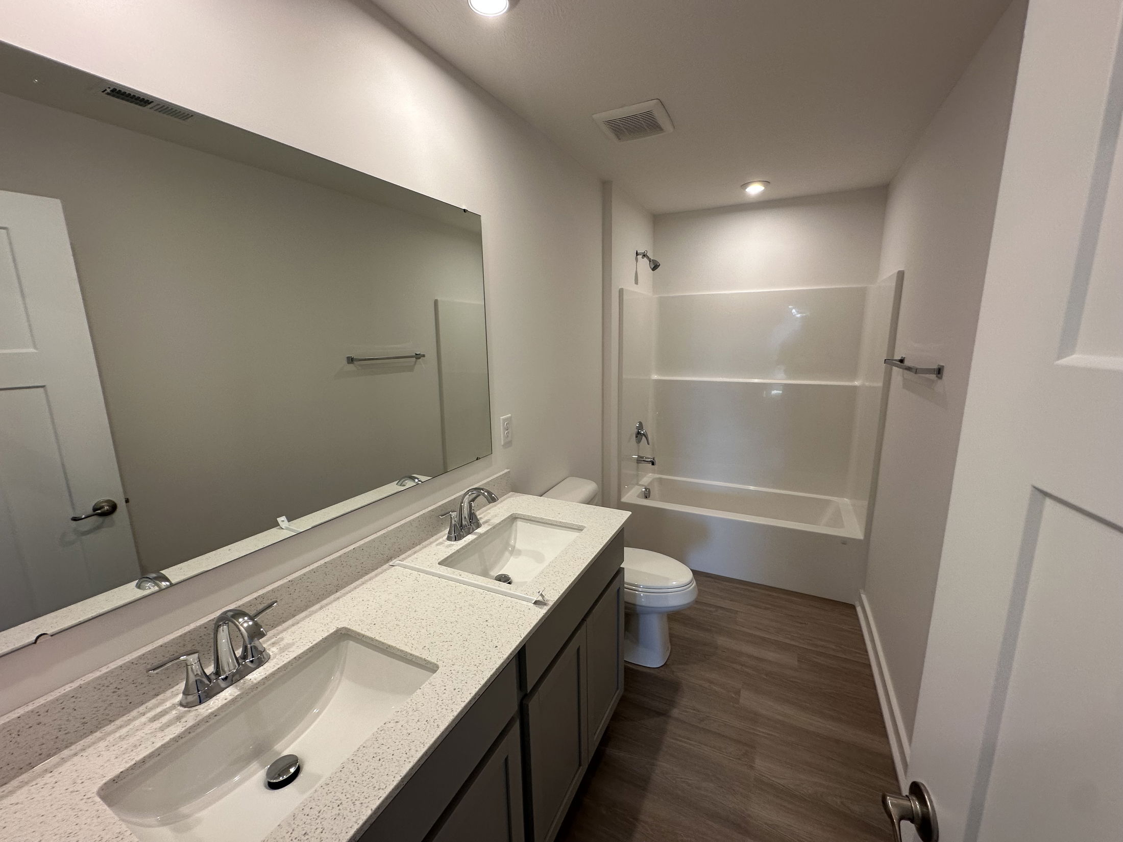 Modern bathroom featuring dual sinks with white countertops, a large mirror, and a bathtub with overhead shower on wood-style flooring.