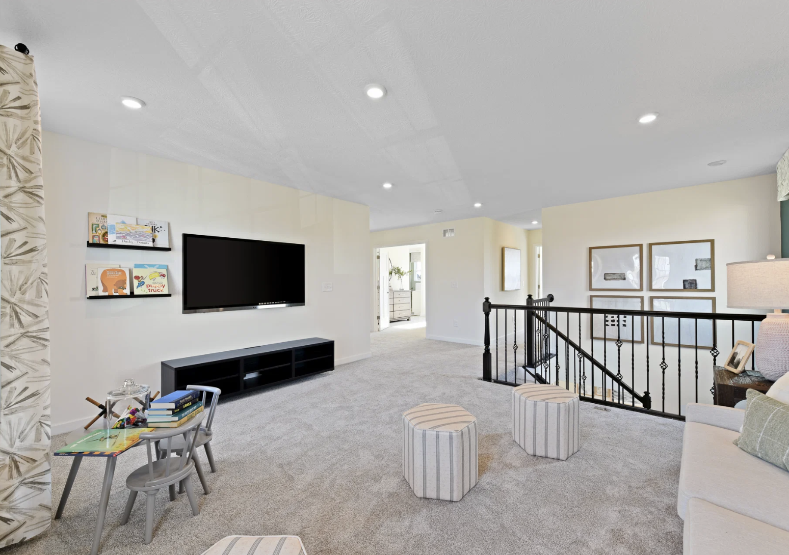 A modern living room featuring a wall-mounted television, light-colored carpet, a sectional sofa, and two striped ottomans, with a table displaying books and decorative items.
