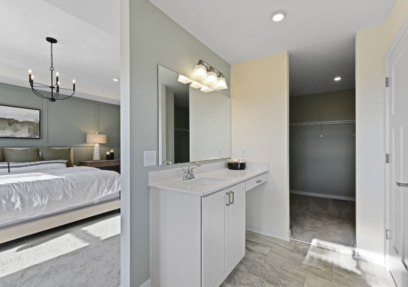 Brightly lit bathroom with a mirror, a white vanity, and a doorway leading to a spacious closet, adjacent to a softly decorated bedroom.