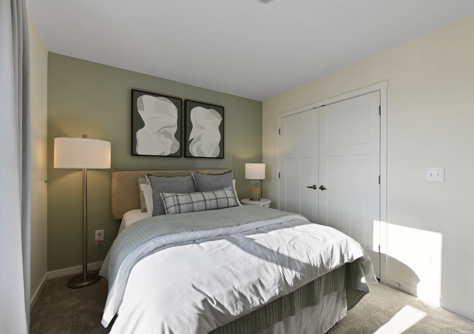 A cozy bedroom featuring a bed with gray and white bedding, two abstract wall art pieces, a floor lamp, and a closet with white double doors.
