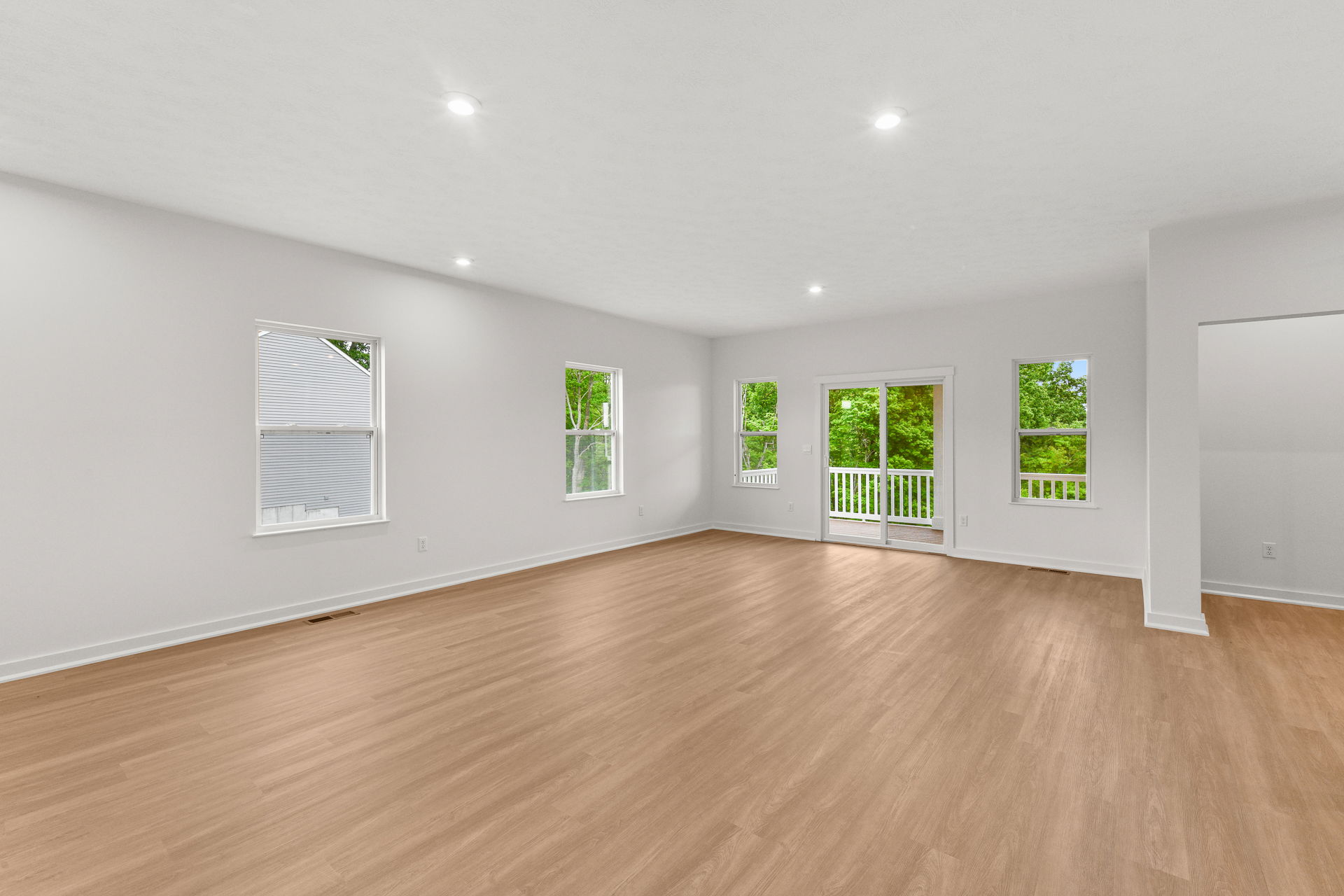 Spacious, modern living room with light wooden flooring, bright windows, and sliding glass doors opening to a balcony with lush green views.