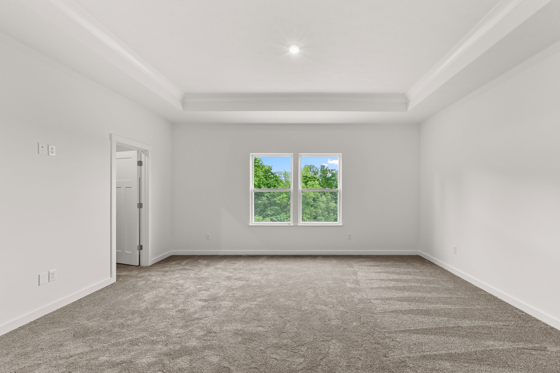 Bright, empty room with beige carpet, white walls, tray ceiling, and a window overlooking green trees.