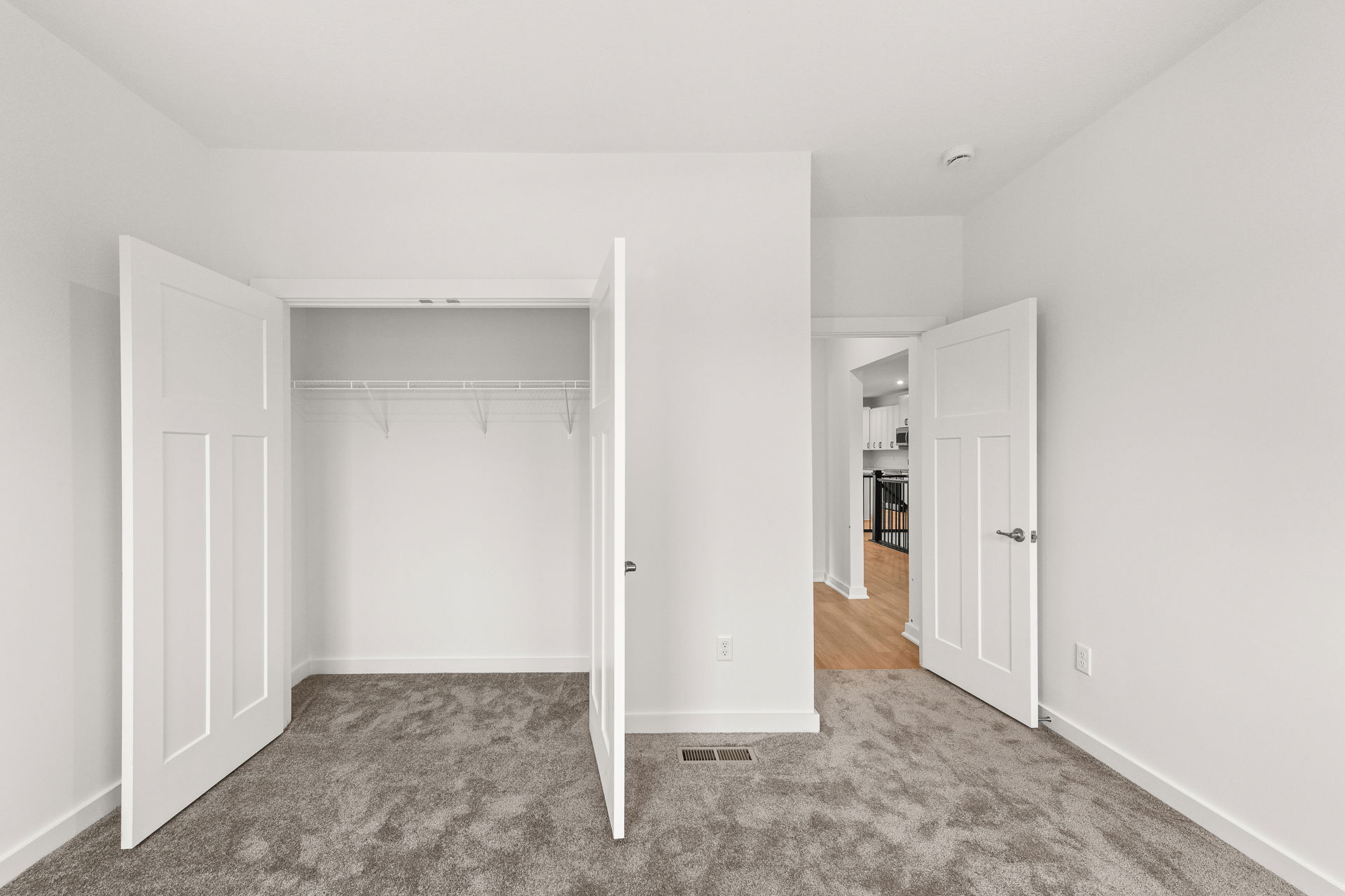 Empty room with an open closet featuring white doors, gray carpet, and hallway view leading to a modern kitchen.
