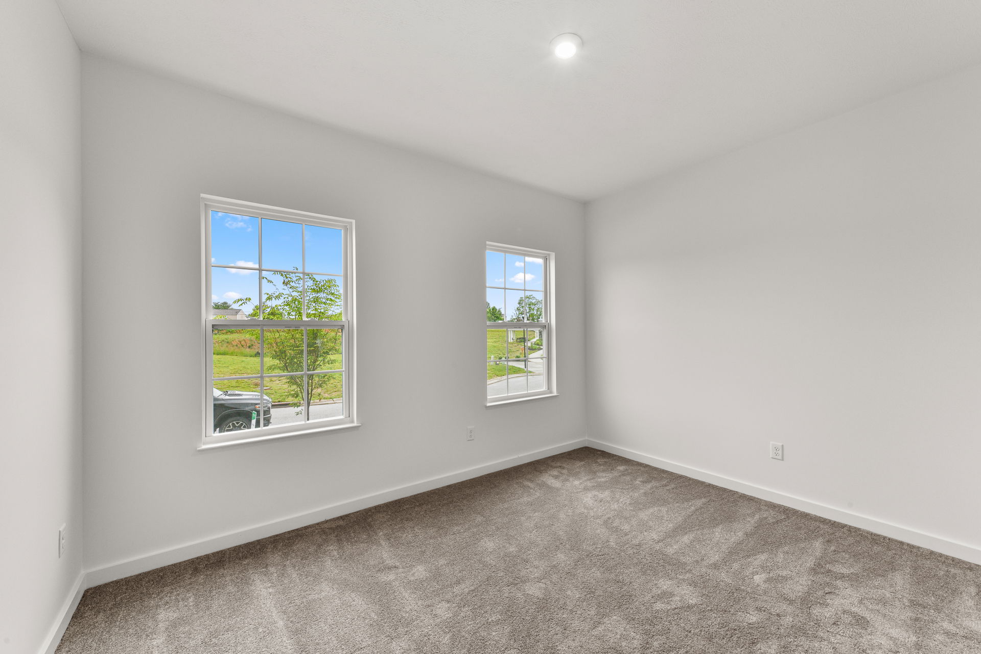 Bright empty room with two large windows, gray carpet, and white walls showcasing scenic outdoor views.