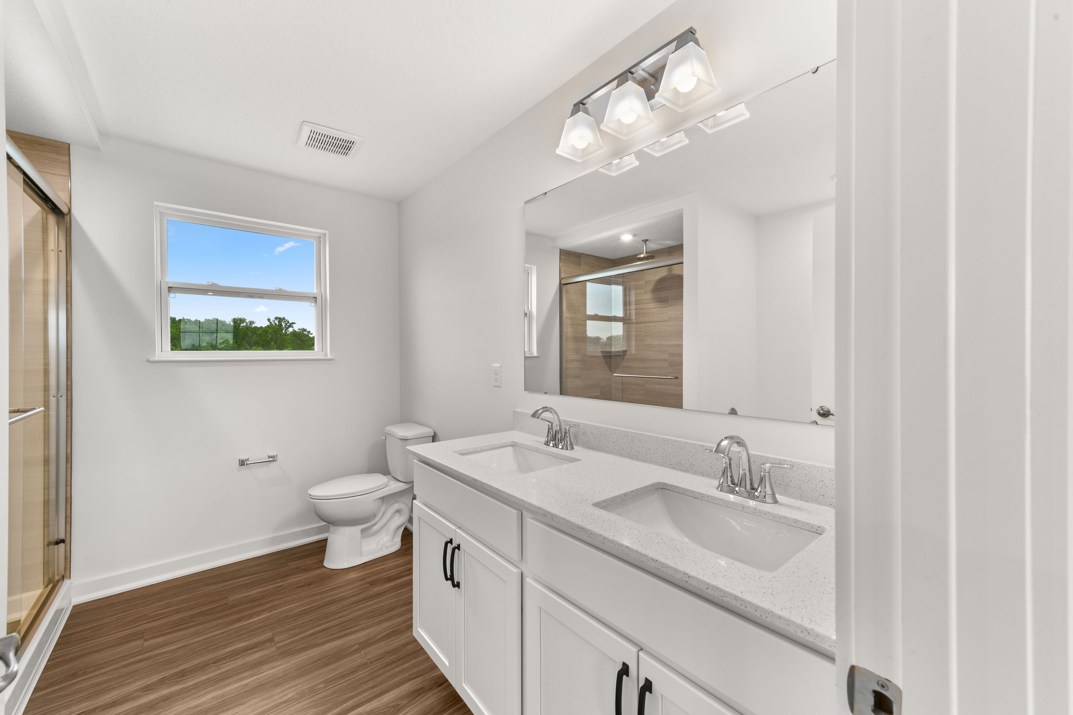 Modern bathroom with dual sinks, white cabinets, wood flooring, and a glass-enclosed shower.