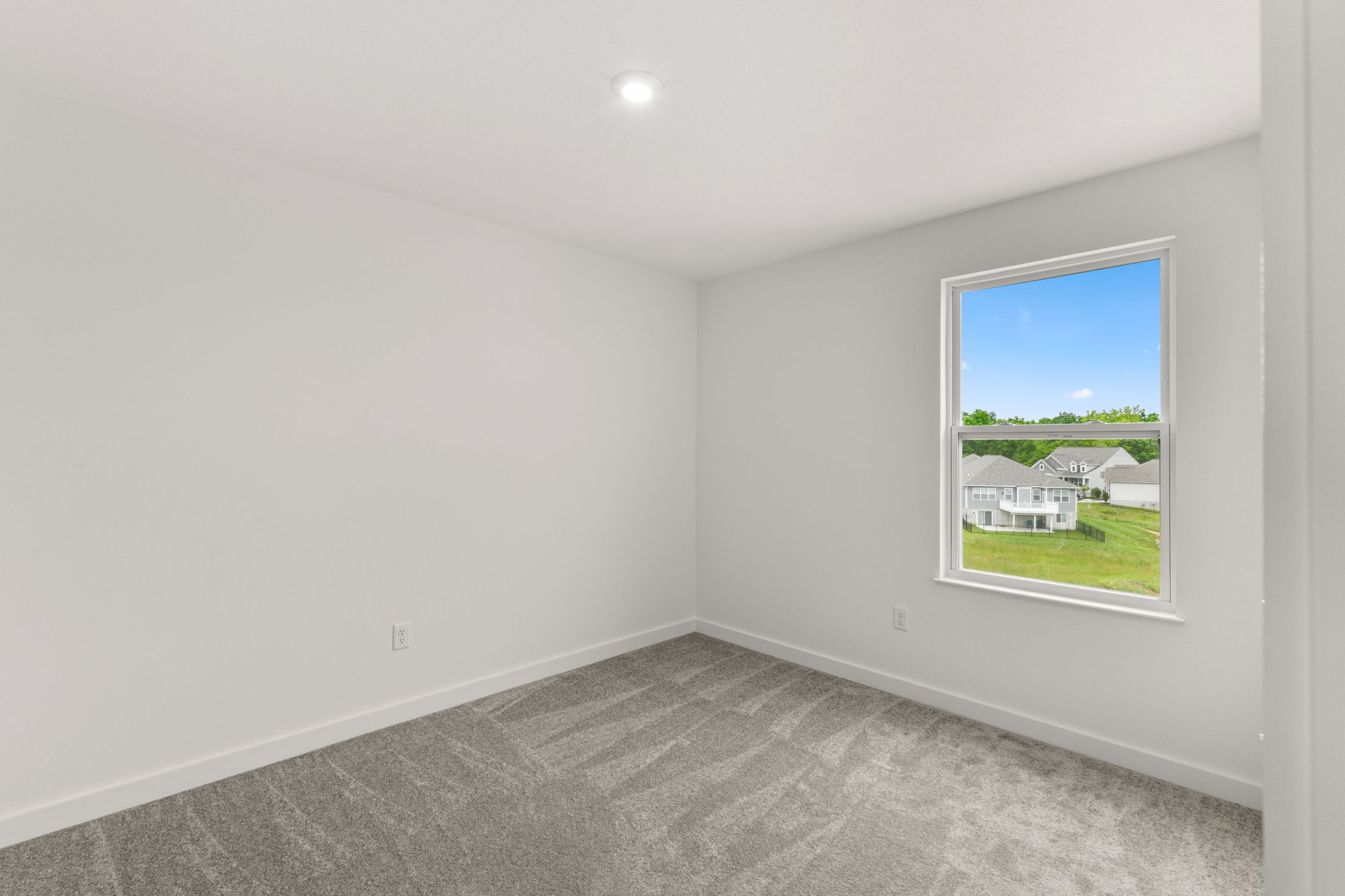 Empty white-walled room with a single window overlooking a suburban neighborhood and gray carpet flooring.