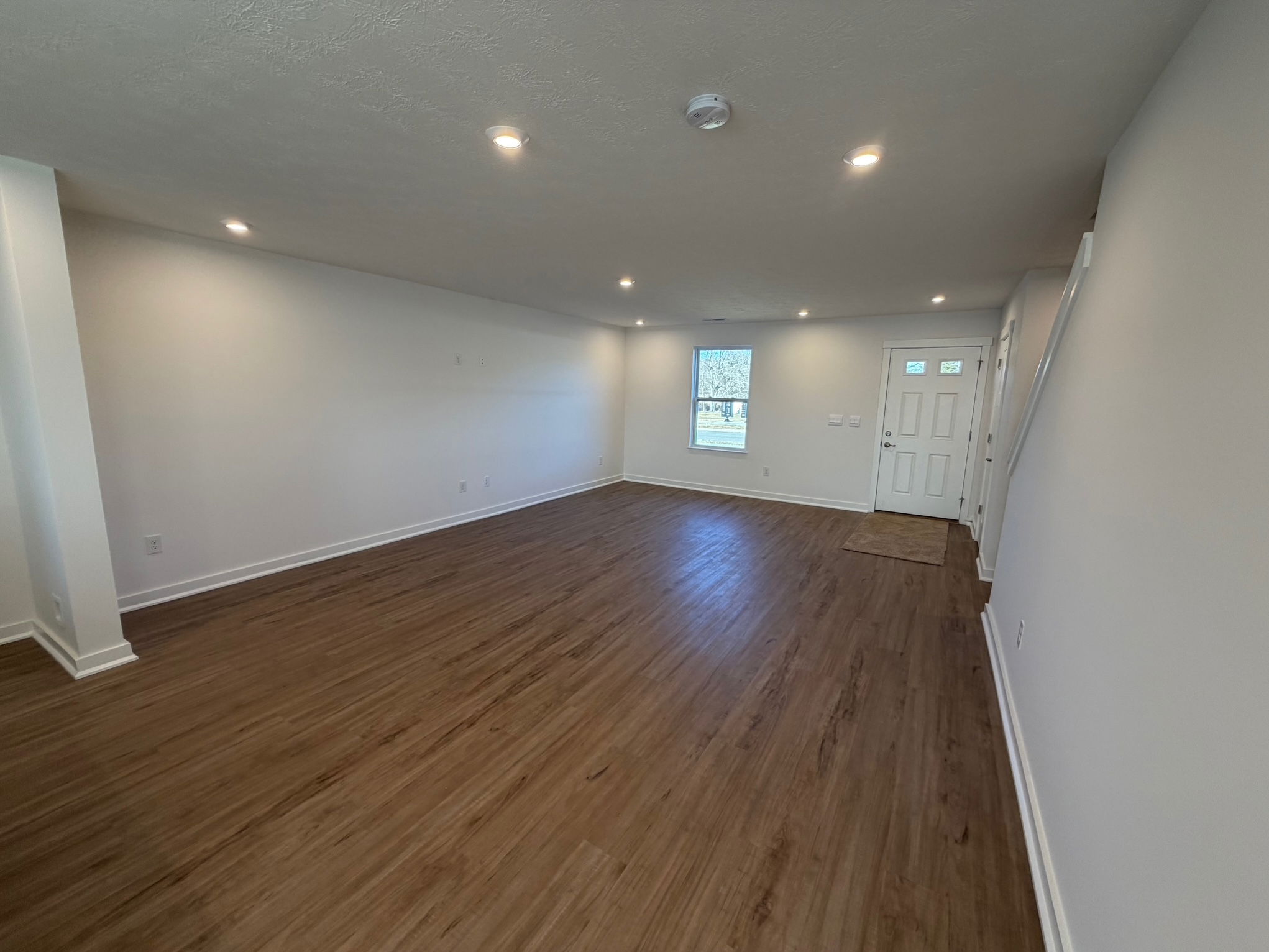 Spacious modern living room with wooden flooring, white walls, and natural light from large window and entrance door.