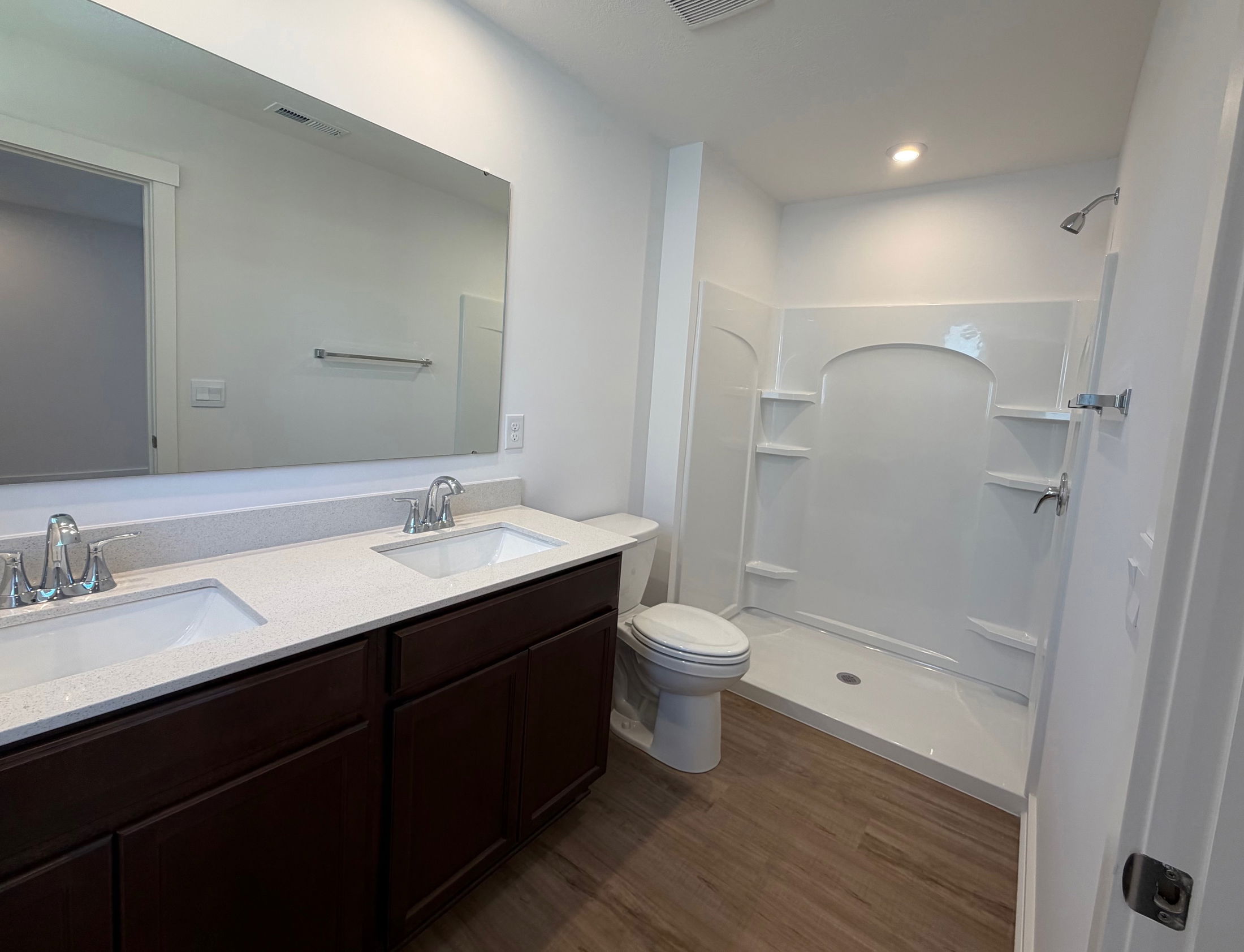 Modern bathroom featuring double sinks, a large mirror, and a spacious shower area with light wood flooring.