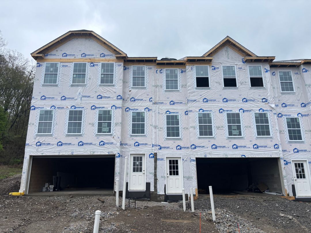 Newly constructed townhouses with protective wrap, featuring multiple windows and garages, under a cloudy sky.