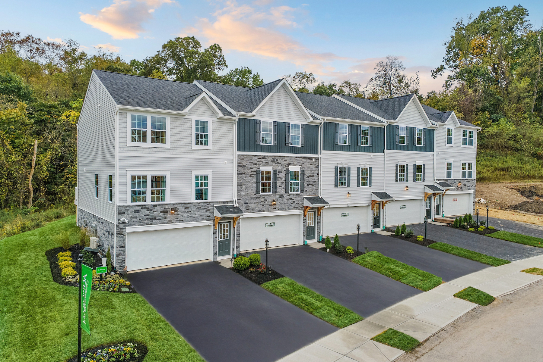Newly constructed townhomes with stone facades and garage doors, surrounded by landscaped greenery and located in a suburban setting.