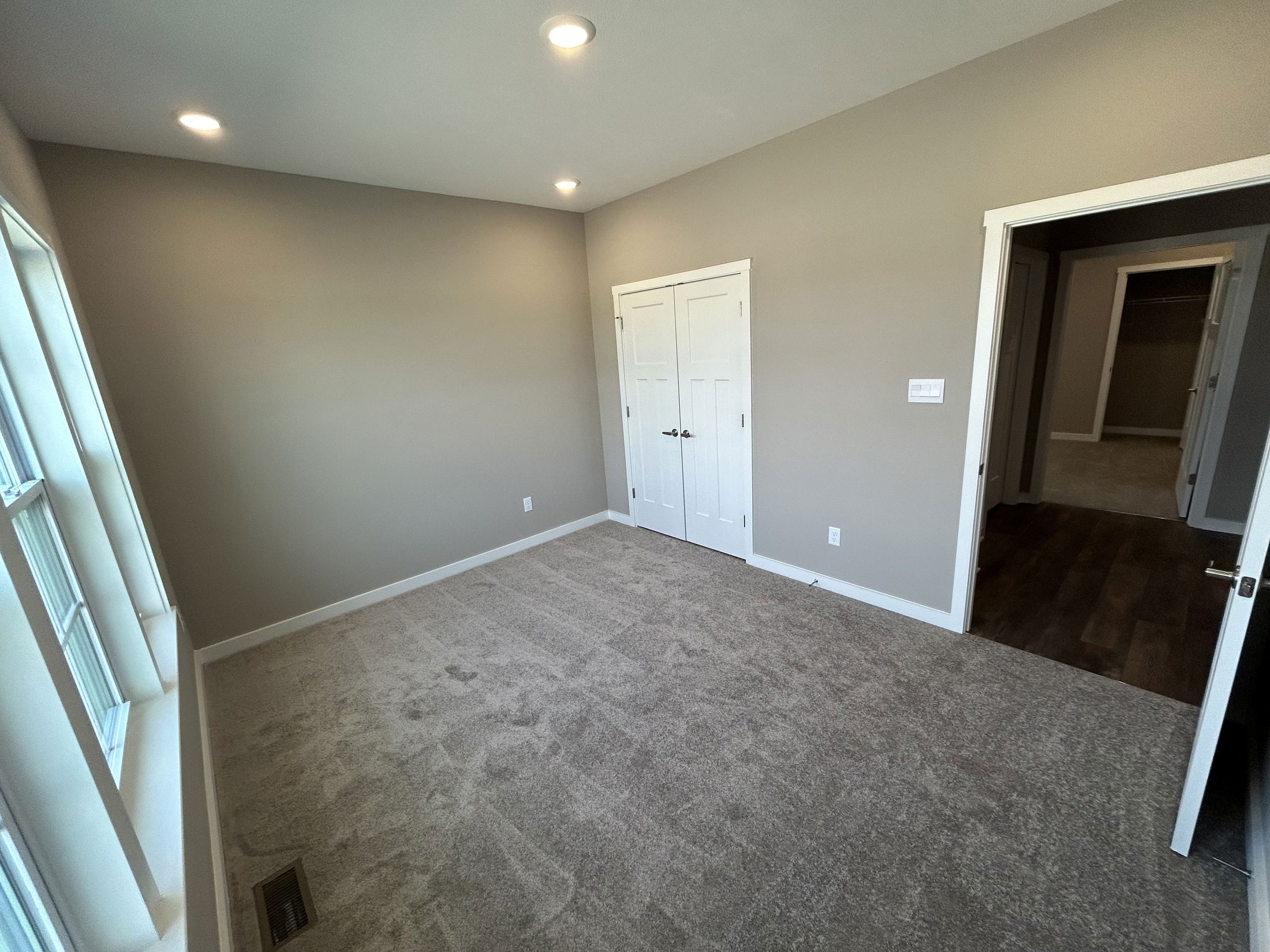 Spacious empty bedroom with beige walls, carpeted floor, recessed lighting, two white double doors, and a large window.