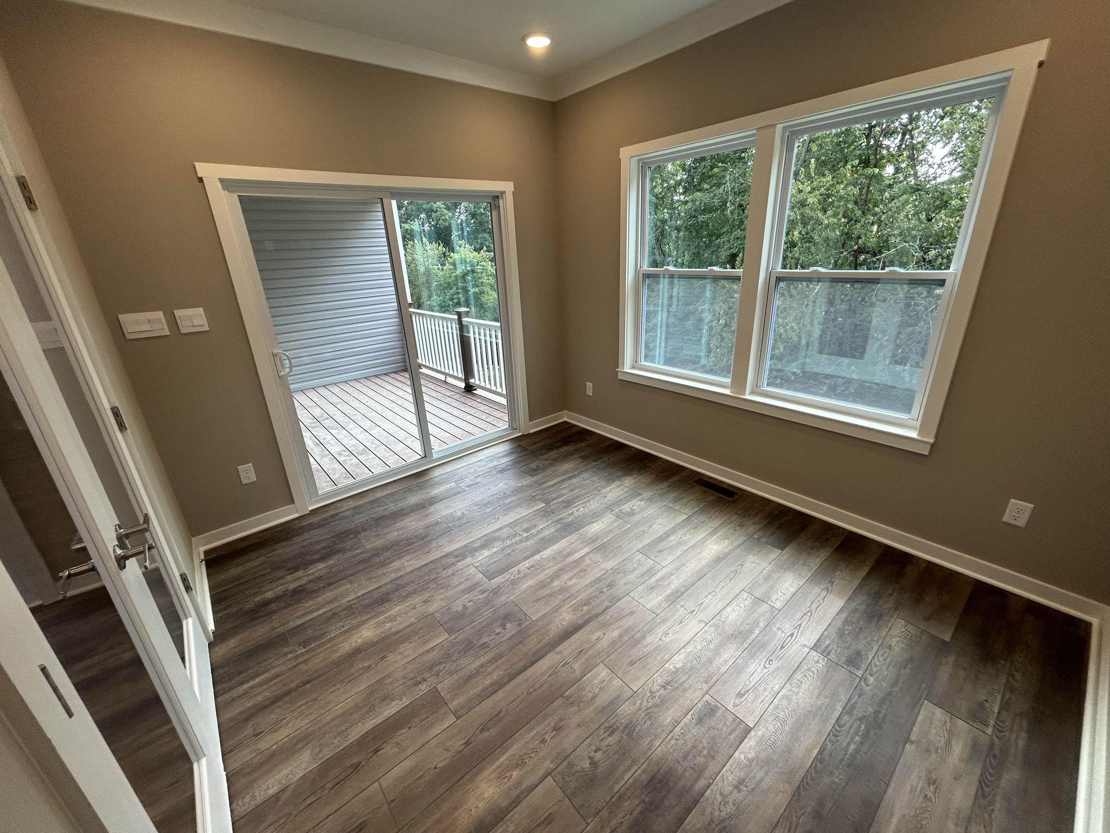 Empty room with wood flooring, large windows, and a sliding glass door leading to a deck with forest views.