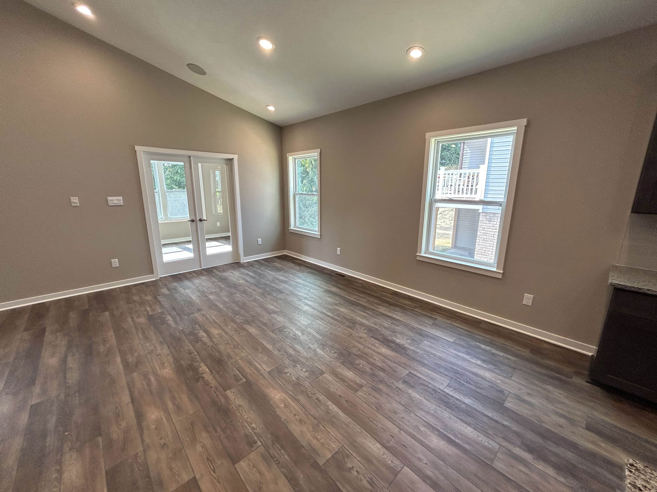 Modern empty living room with wood flooring, beige walls, recessed lighting, and French doors leading to another room.