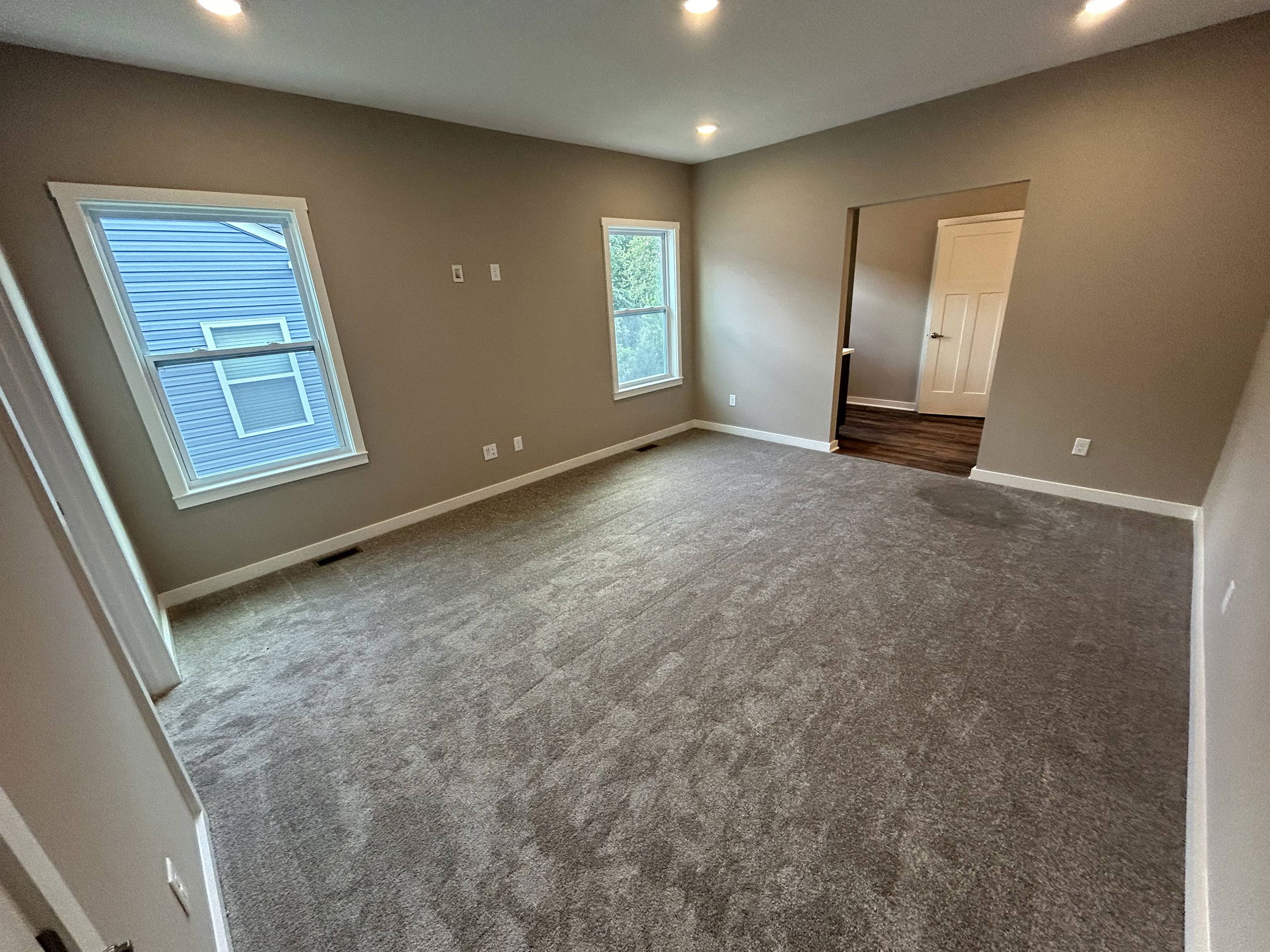 Empty carpeted room with beige walls, two windows, and an entrance to a hallway.