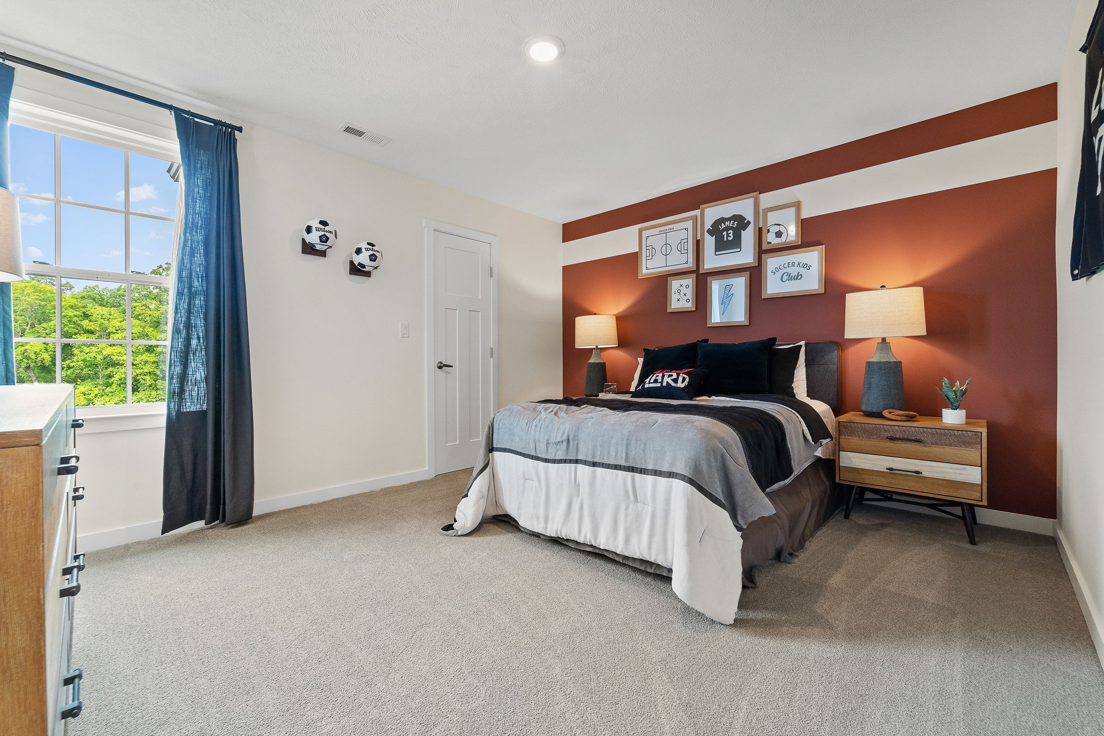 A modern bedroom with soccer-themed decor, featuring a bed against a red accent wall with framed art, two bedside lamps, and a window with a view of greenery.
