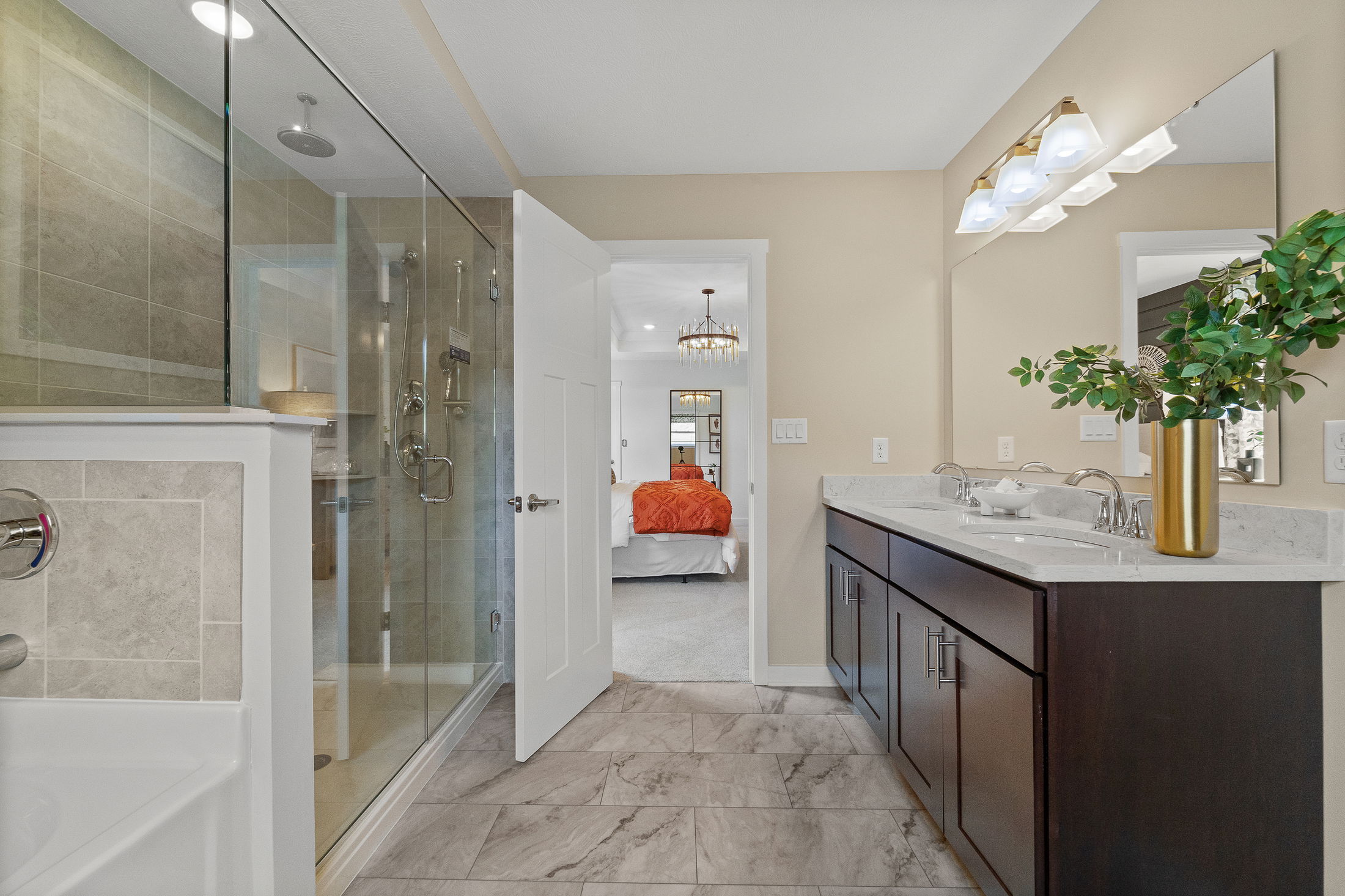 Modern bathroom with a glass-enclosed shower, dual sink vanity, marble flooring, and view into a bedroom with an orange bedspread.