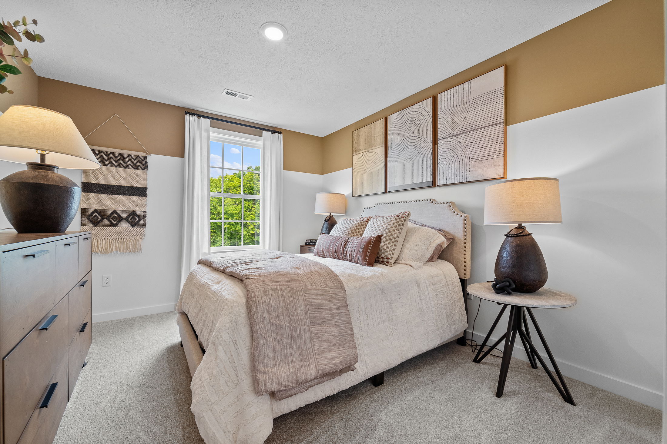 Modern bedroom with neutral tones, featuring a cozy bed, decorative pillows, artistic wall decor, natural light from a window, and stylish lamps.