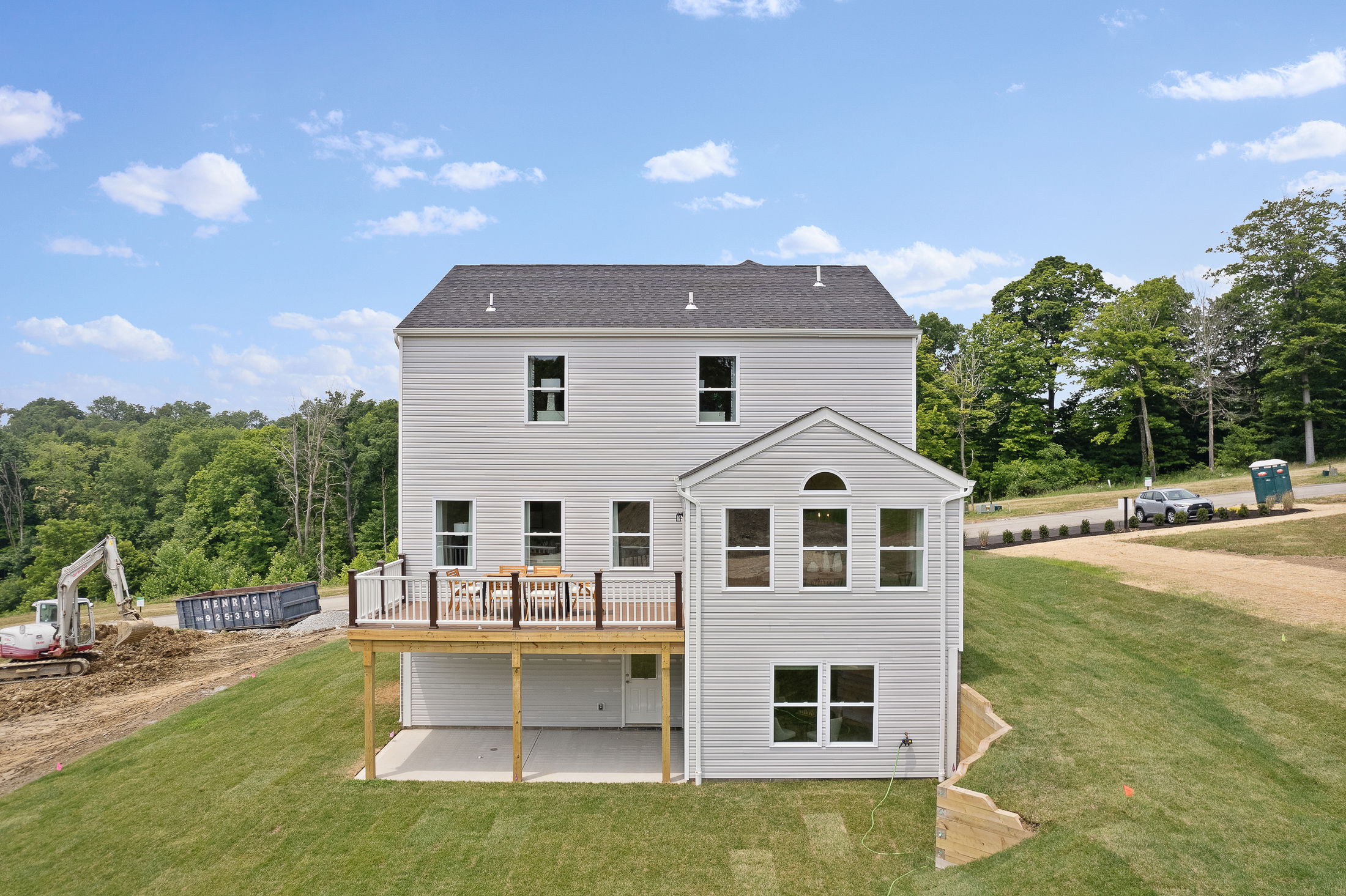Rear view of a modern two-story house with a wooden deck, under construction, surrounded by lush greenery.