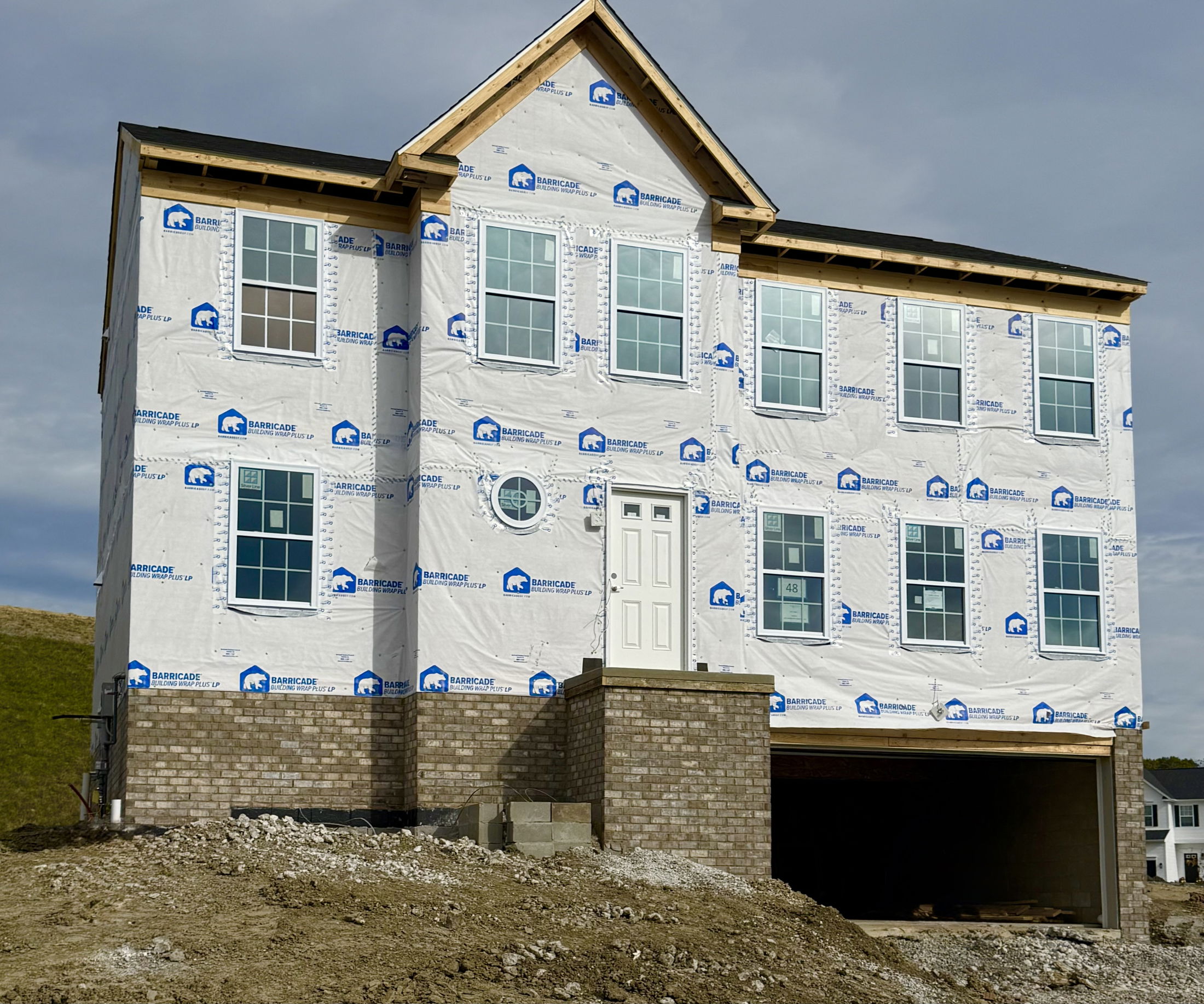 New two-story house under construction with visible Barricade building wrap and garage, situated on a dirt lot.