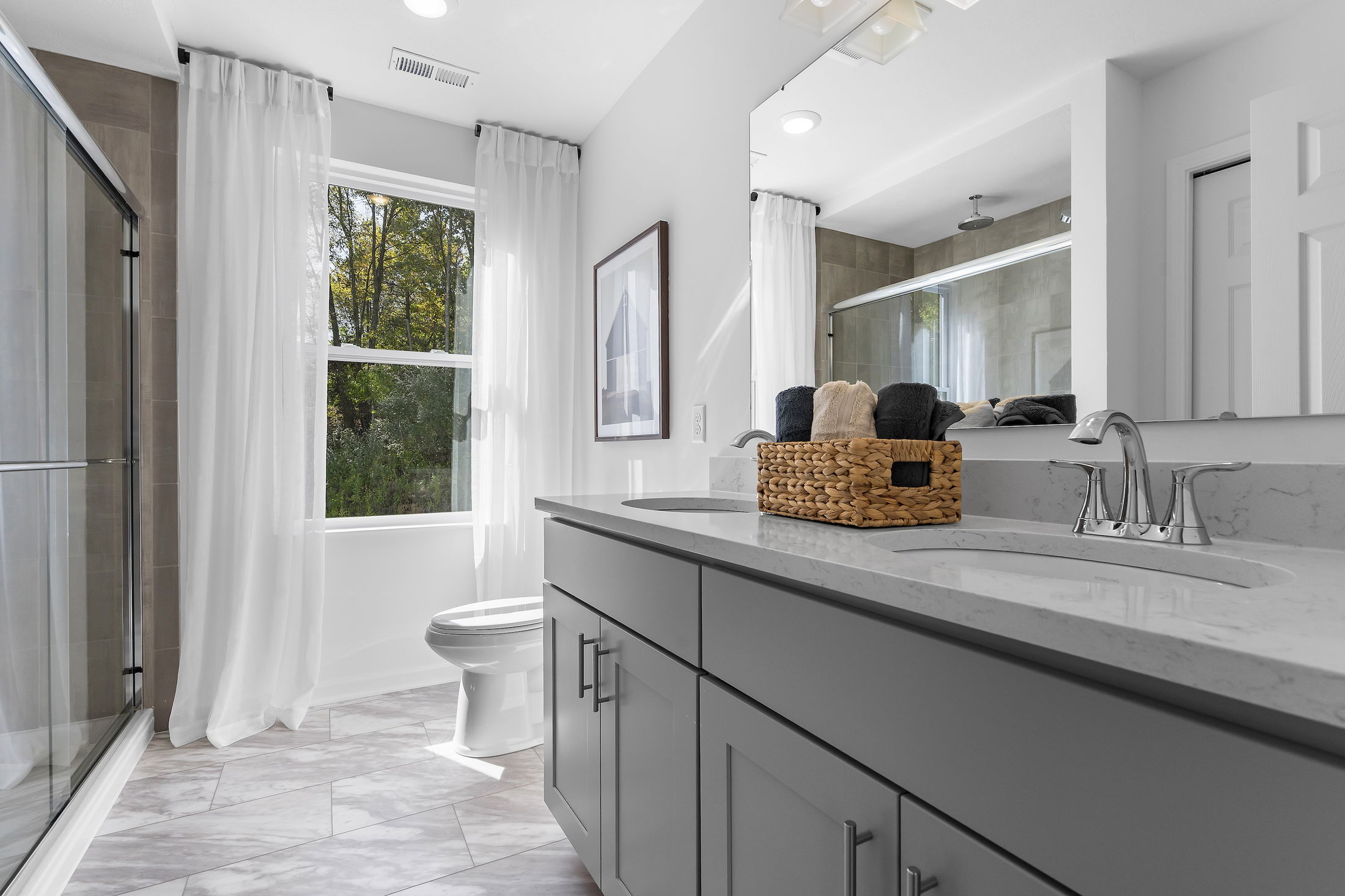 Modern bathroom with gray cabinetry, marble countertop, large mirror, wicker basket, glass shower, and a window with sheer curtains.