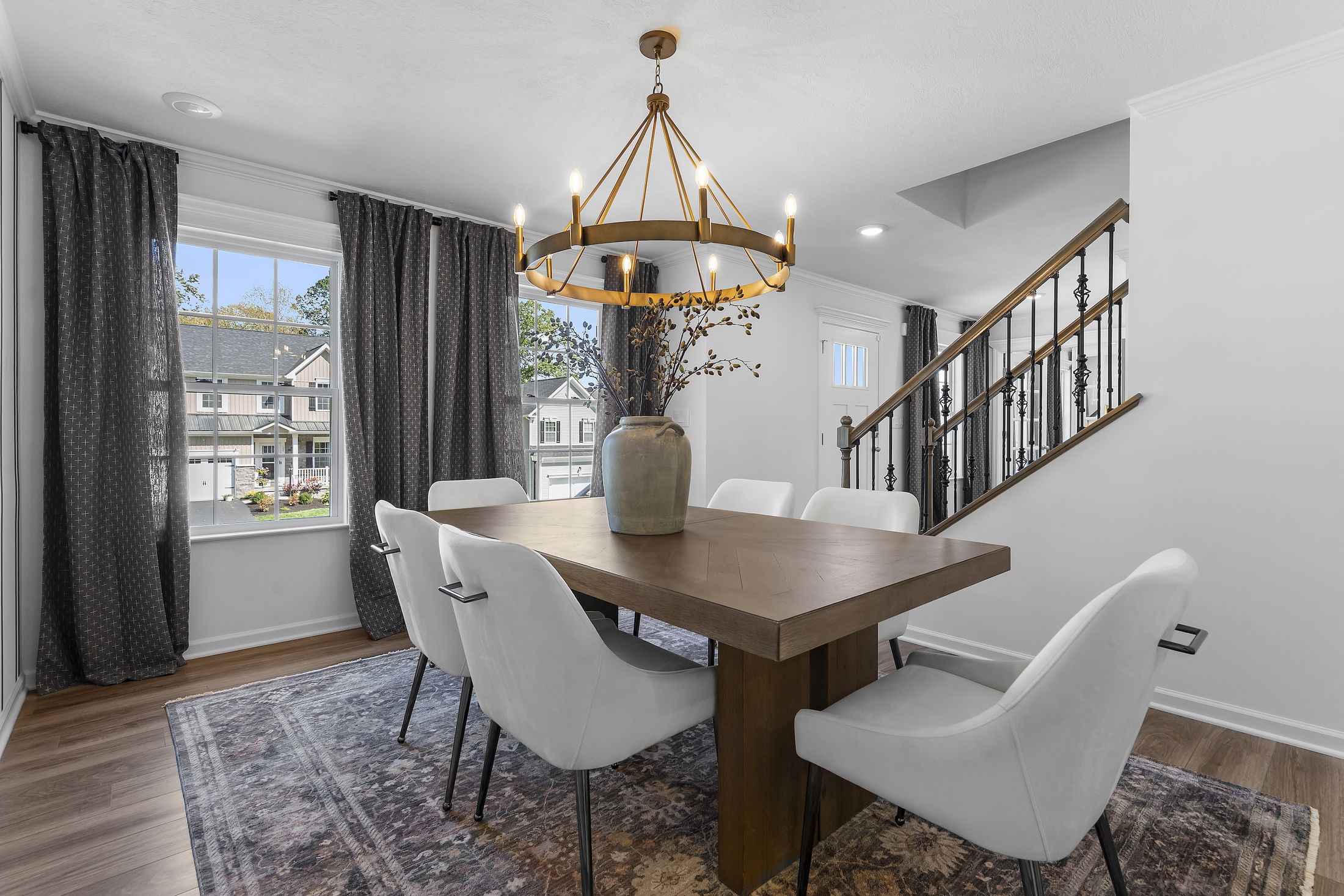 Stylish dining room with a modern chandelier, wooden table, white chairs, and decorative vase, overlooking residential neighborhood through large window.