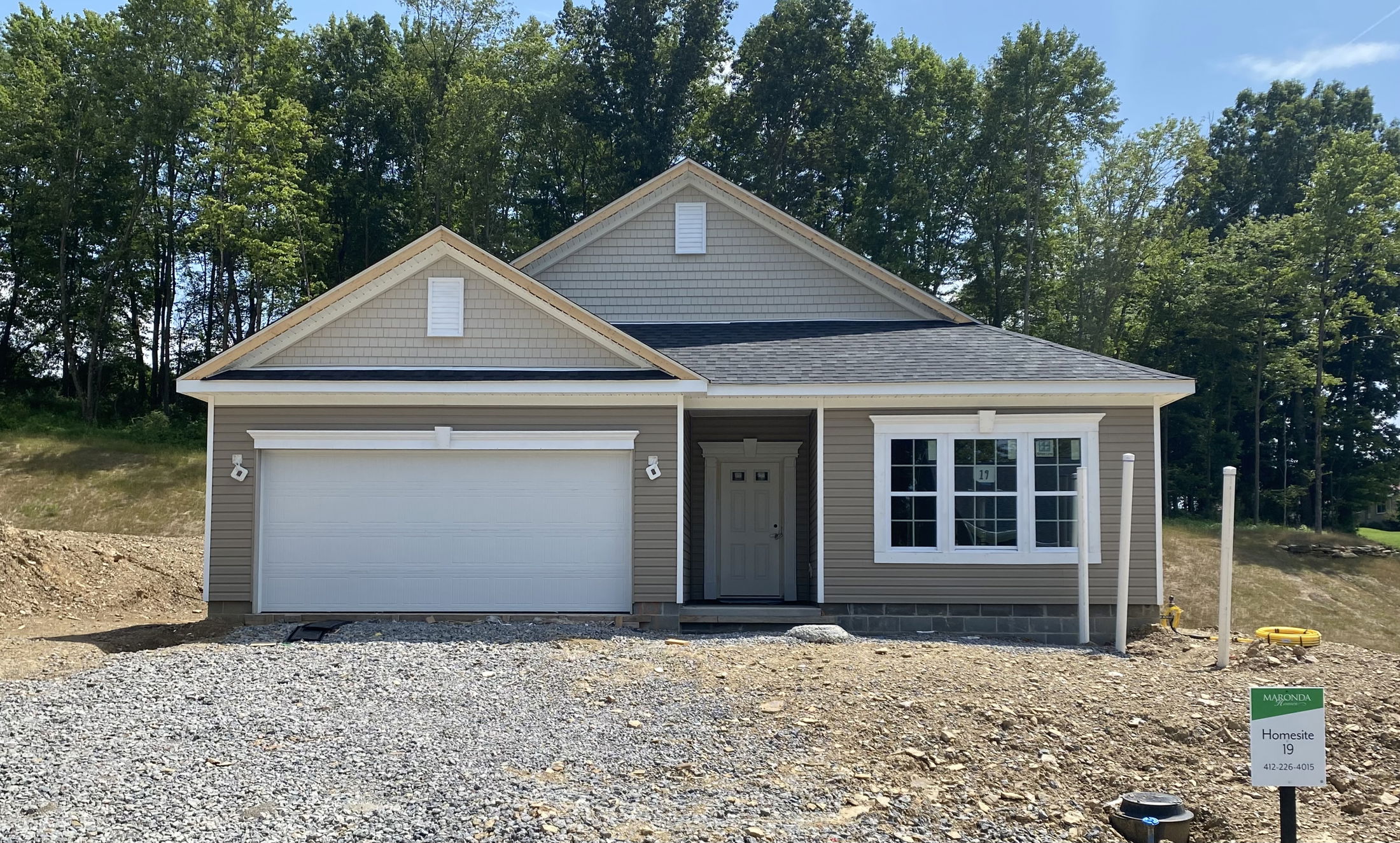 Newly constructed single-story home with a two-car garage and tan siding, surrounded by trees.
