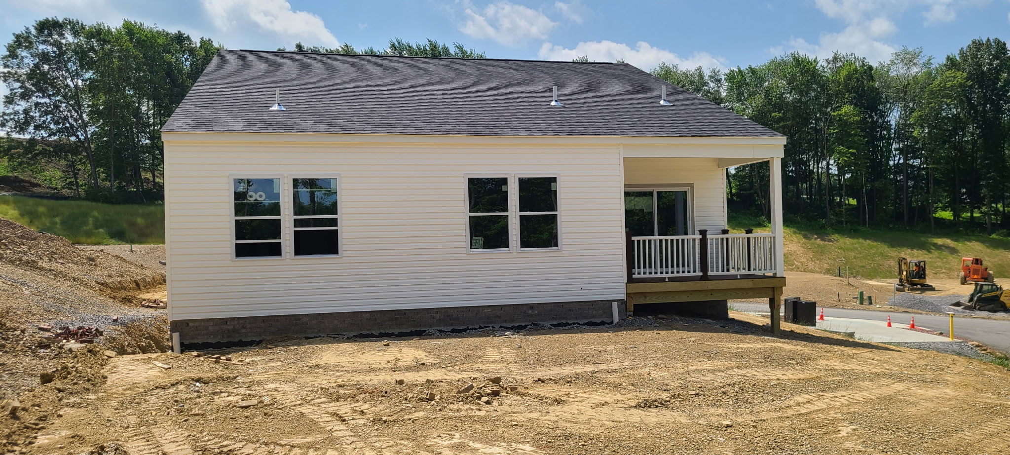 A newly constructed house with white siding and a small porch, surrounded by dirt and construction vehicles under a clear blue sky.