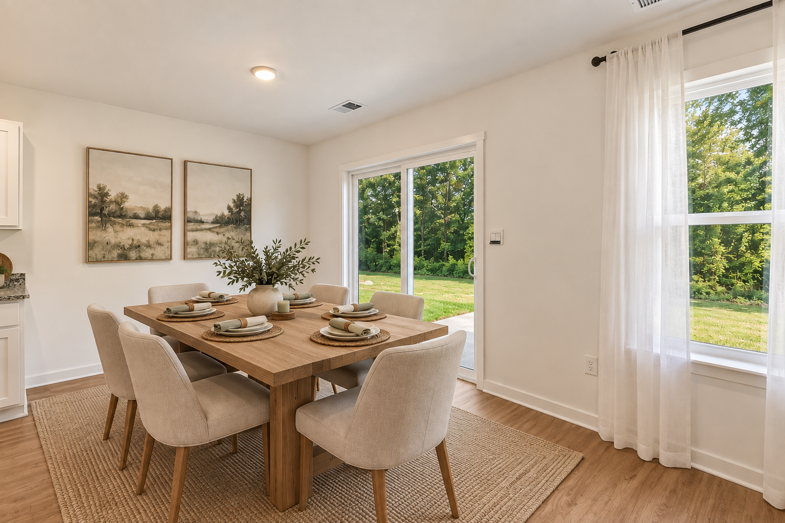 A modern dining room featuring a wooden table set for six, with beige chairs and artwork on the walls, overlooking a green yard through glass sliding doors.
