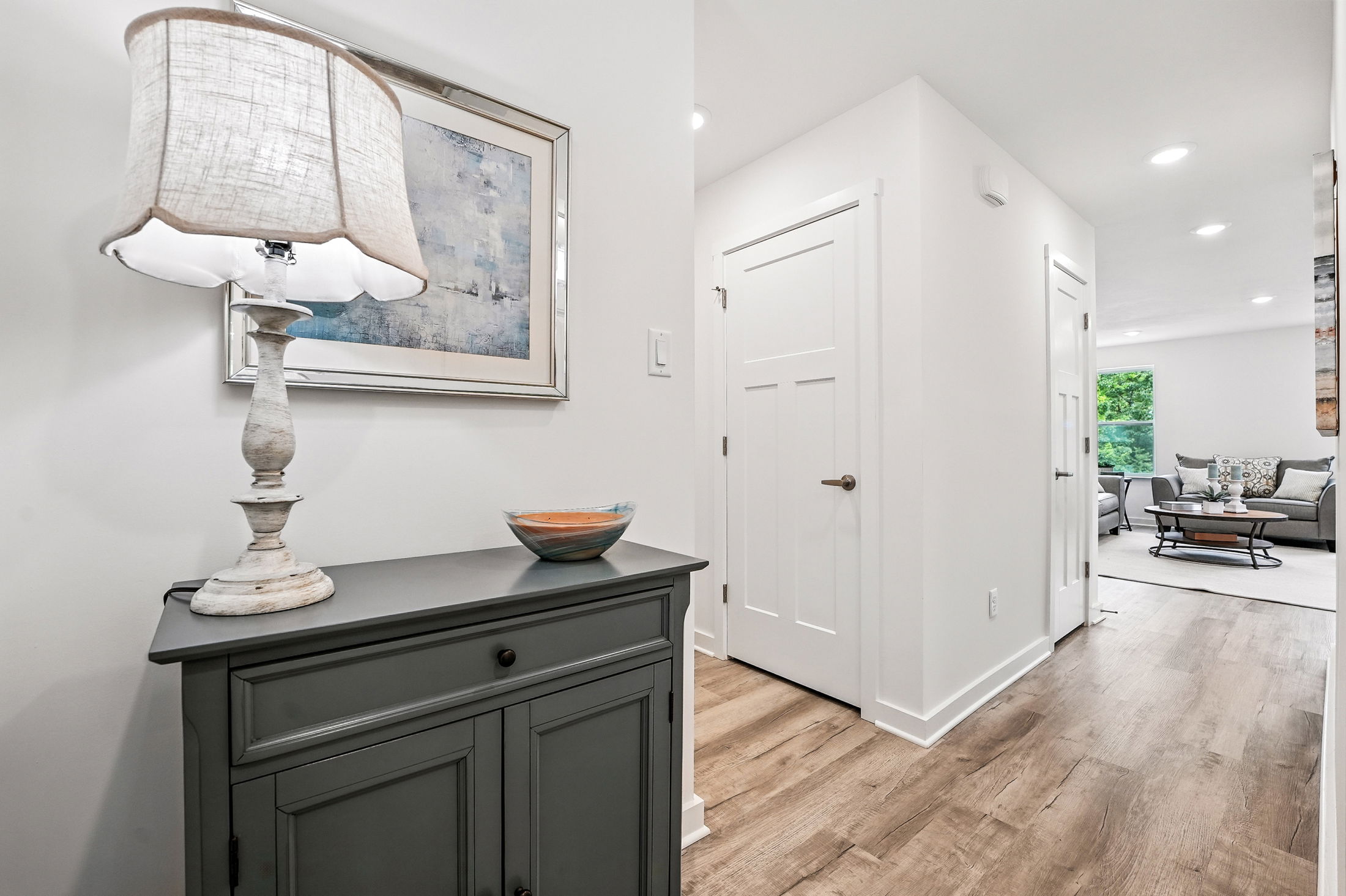 Modern hallway interior with wooden flooring, a gray cabinet, decorative lamp, and abstract wall art leading to a living room area.