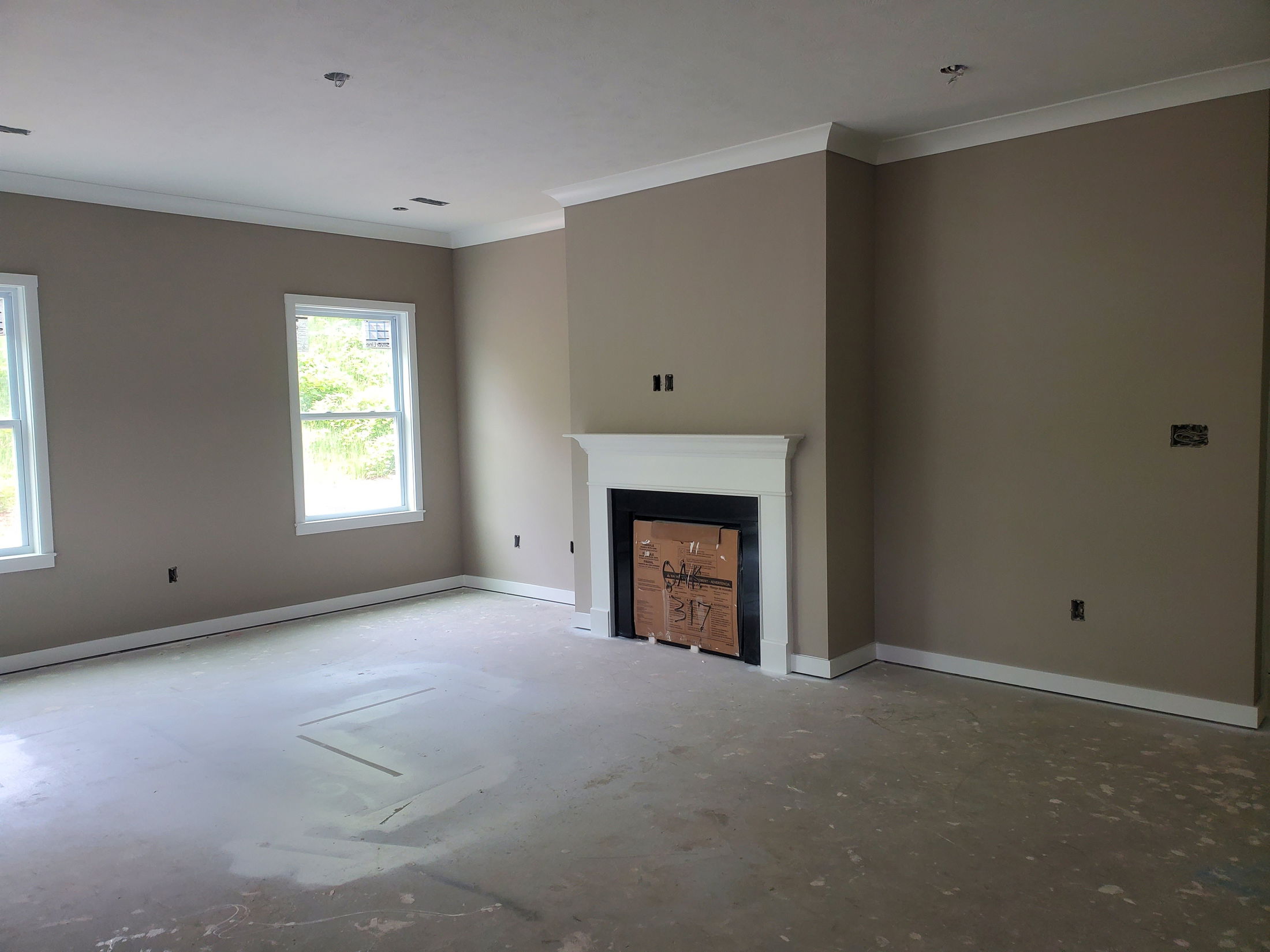 Empty, unfinished living room featuring a fireplace with cardboard protection, beige walls, and two large windows, ready for interior design.