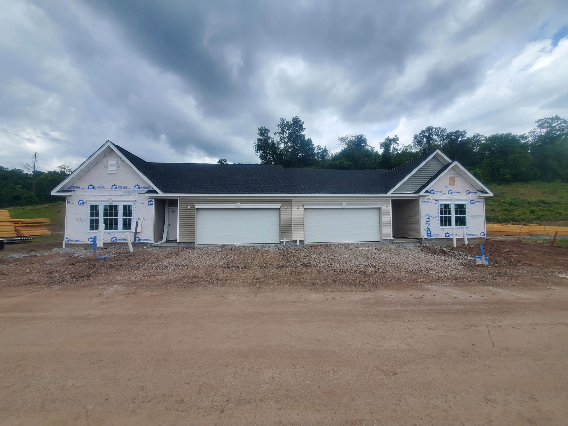 Newly constructed duplex with two garages and siding partially installed, set against a backdrop of cloudy skies and greenery.