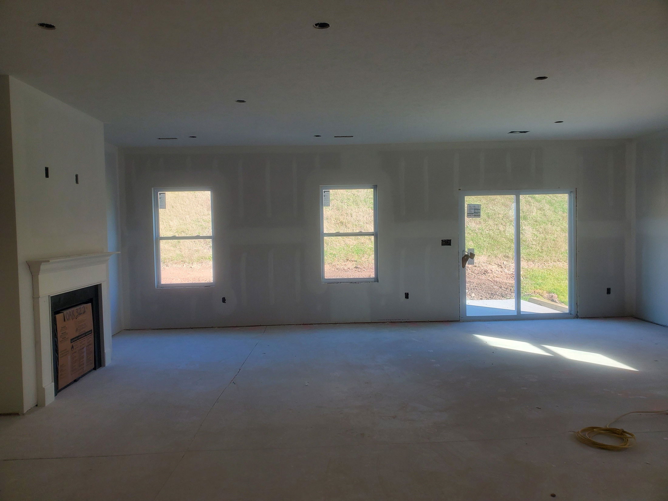 Empty living room under construction with white drywall, three windows, a fireplace, and sliding glass doors leading outside.