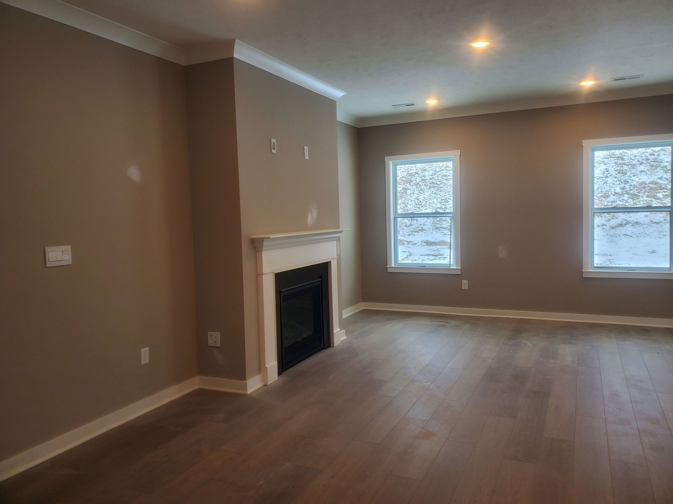 Empty living room with beige walls, wooden flooring, a white-framed fireplace, and two large windows.