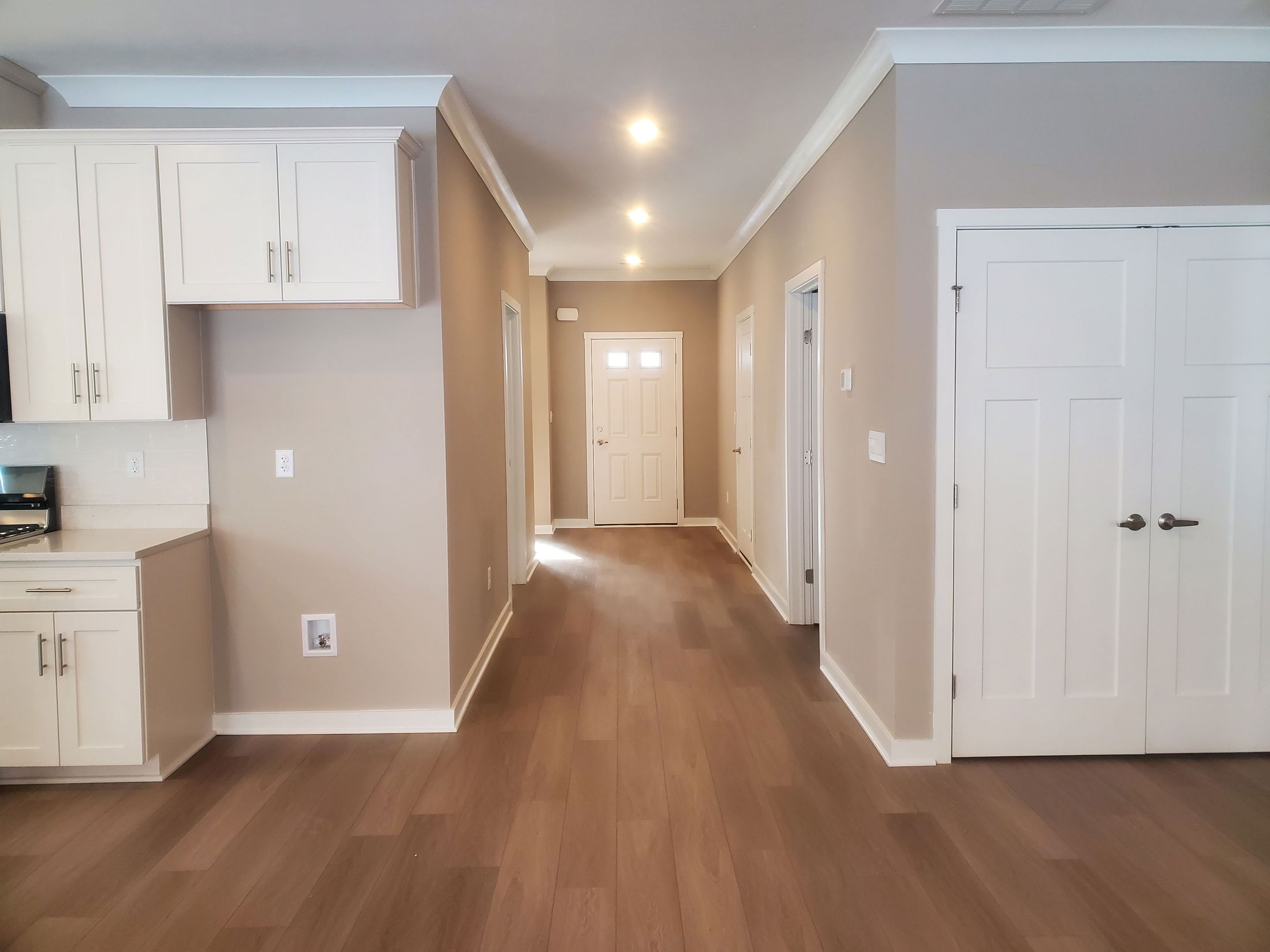 Modern kitchen interior with light wood flooring, beige walls, and natural light illuminating the hallway and entry door.