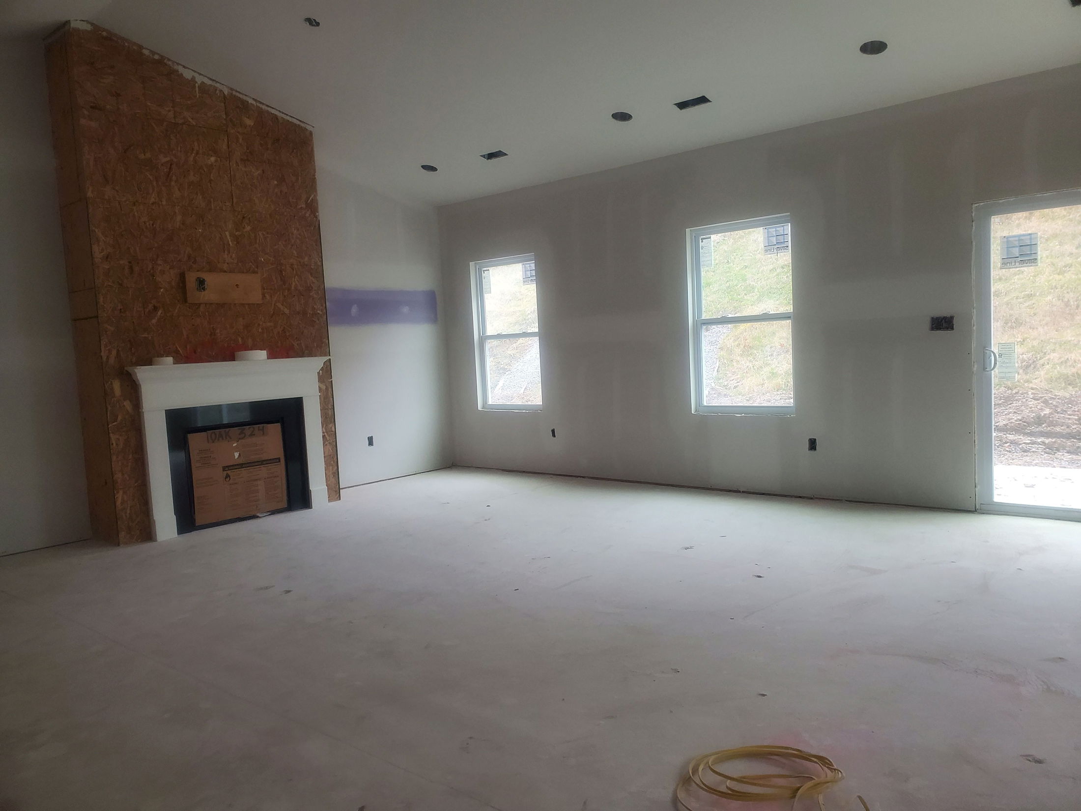 Unfinished living room interior with drywall, a fireplace under construction, and large windows providing natural light.