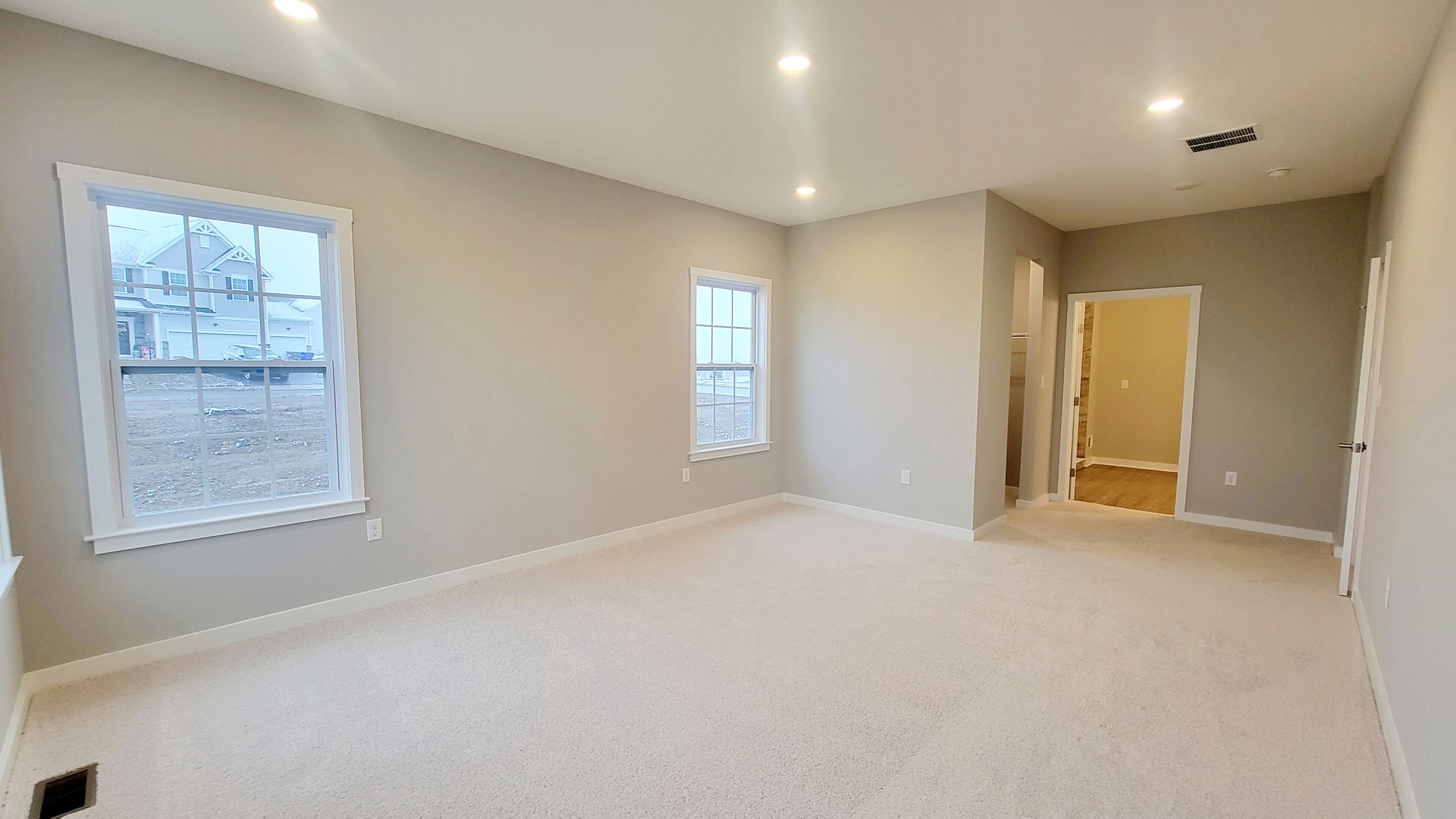 Spacious beige carpeted bedroom with gray walls, recessed lighting, large windows, and an open doorway to a wooden floored area.