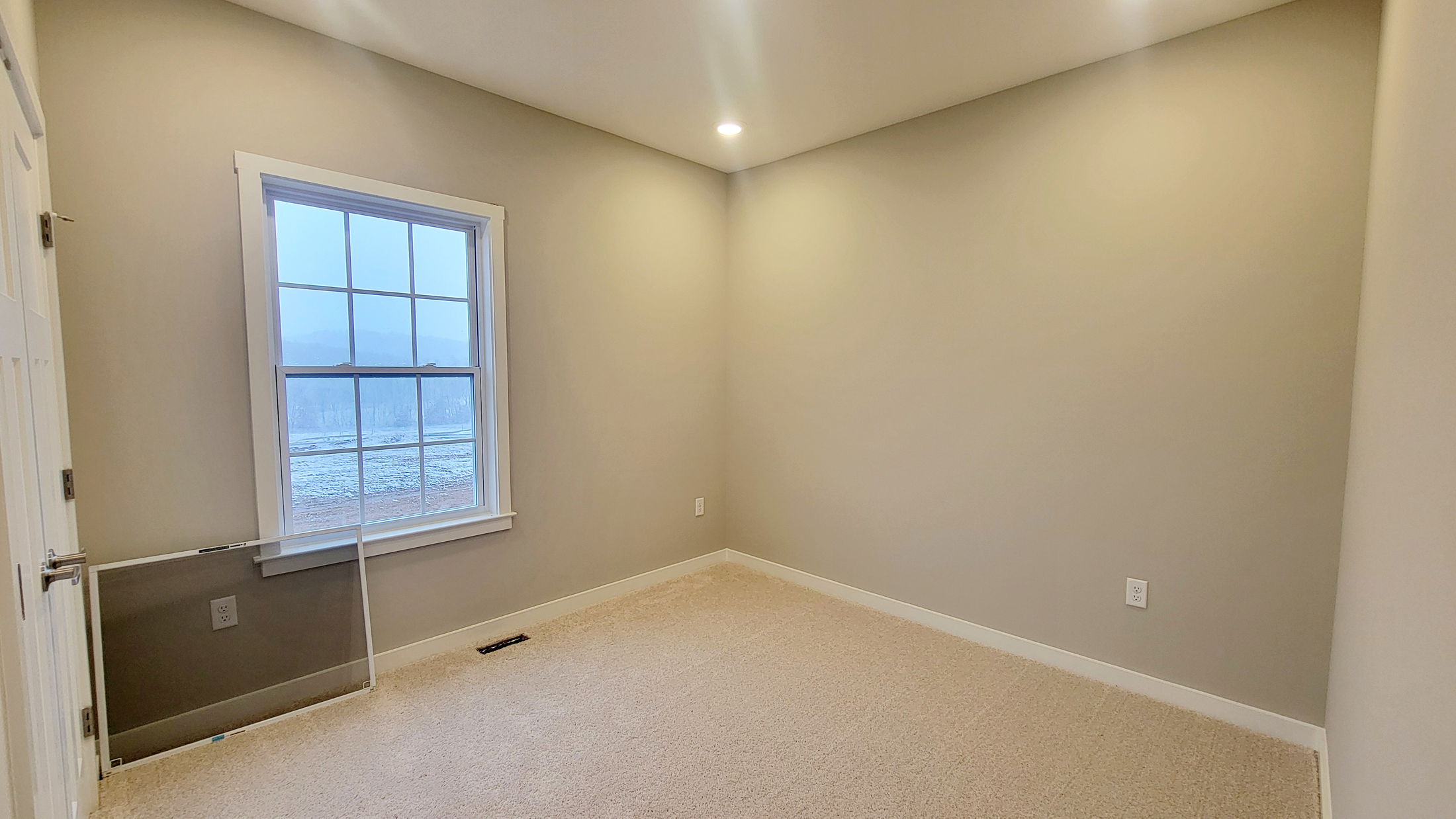 Empty beige room with carpet, recessed lighting, and a large window overlooking a snowy landscape.