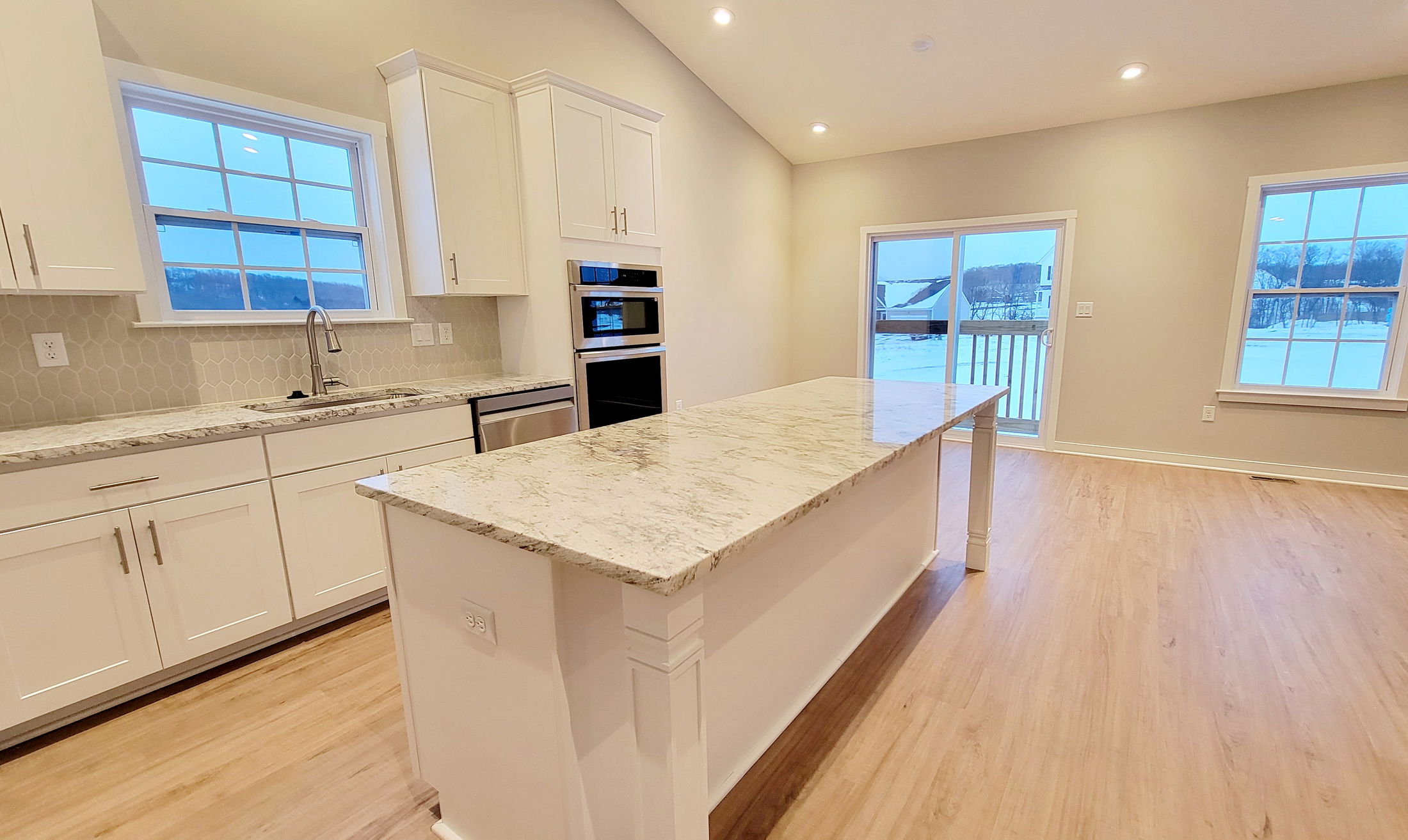A modern kitchen with white cabinetry, gray countertops, stainless steel appliances, and wood flooring, featuring large windows and recessed lighting.