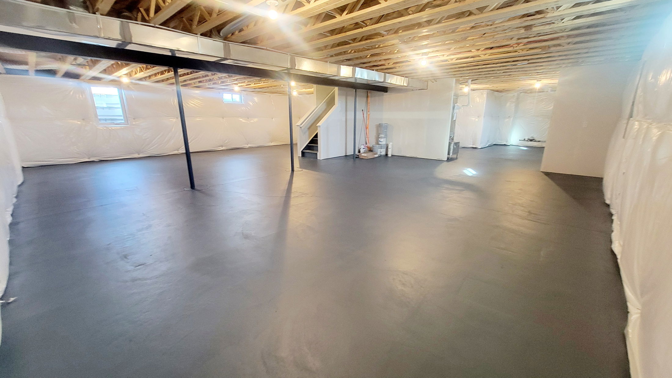 Spacious unfinished basement with exposed wooden beams and concrete floor, featuring small windows and a staircase.
