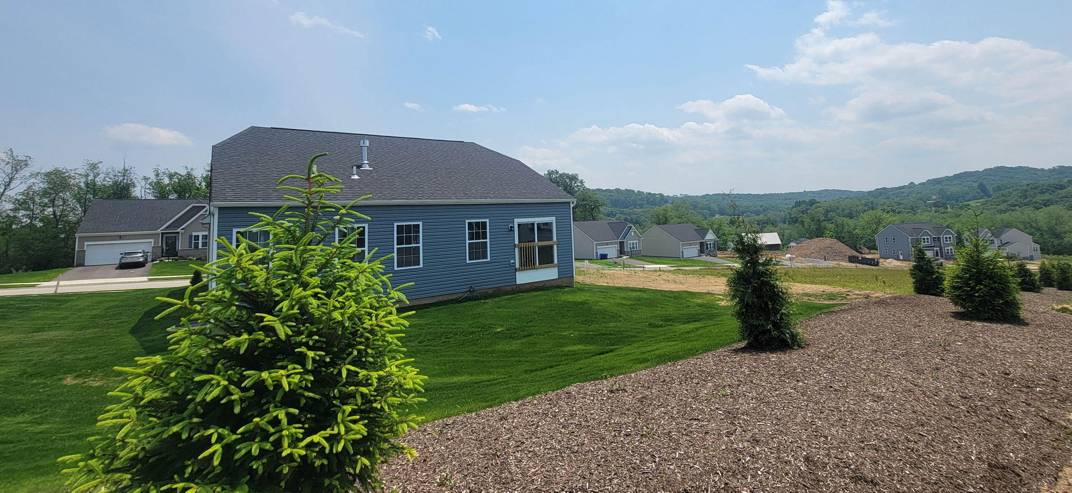 Suburban landscape featuring a blue house with a well-maintained lawn, small trees, and rolling hills in the background under a clear sky.