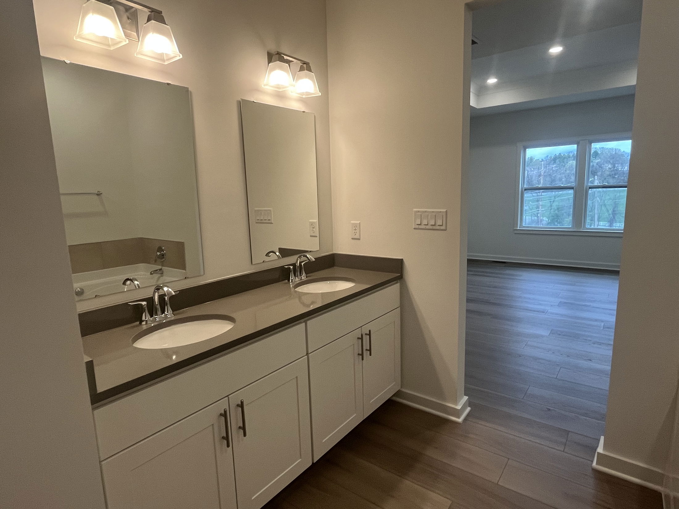 Modern bathroom with double sinks, large mirrors, and elegant lighting leading into a spacious room with wood flooring and natural light.